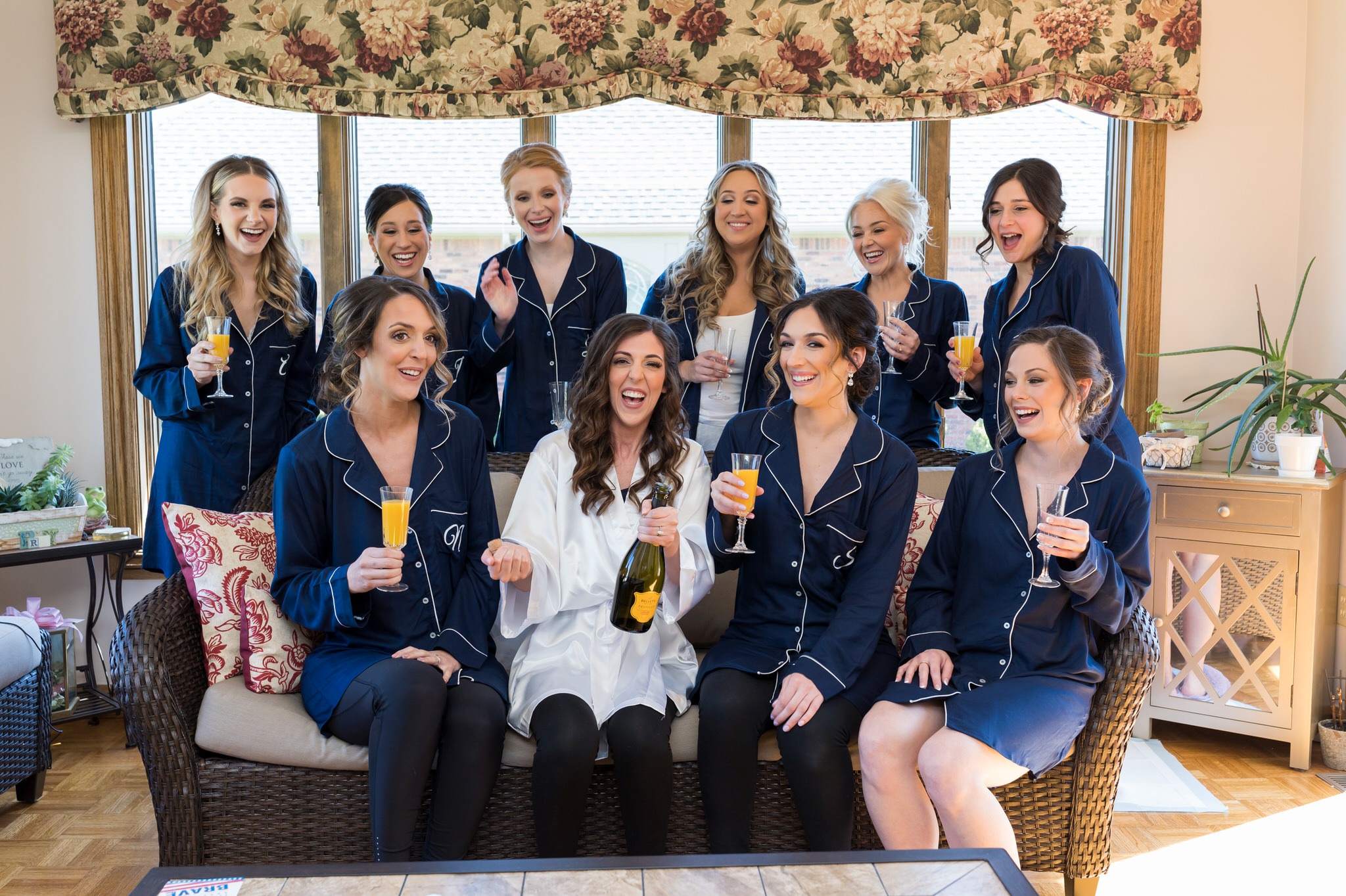 bridesmaids popping champagne wearing matching robes sitting on a wicker bench 