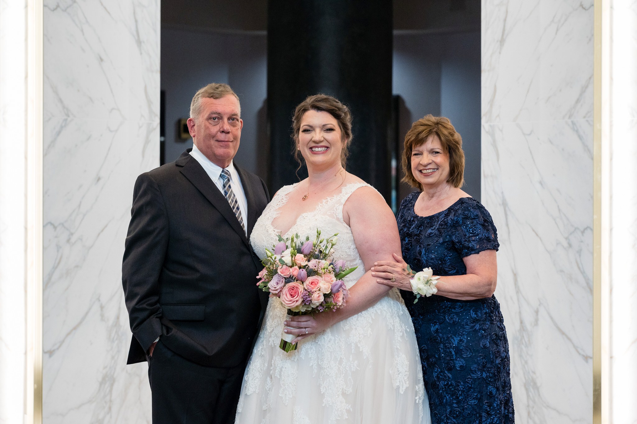 a bride poses for a formal photo with her mom and dad on either side of her at the Atheneum Hotel in Detroit