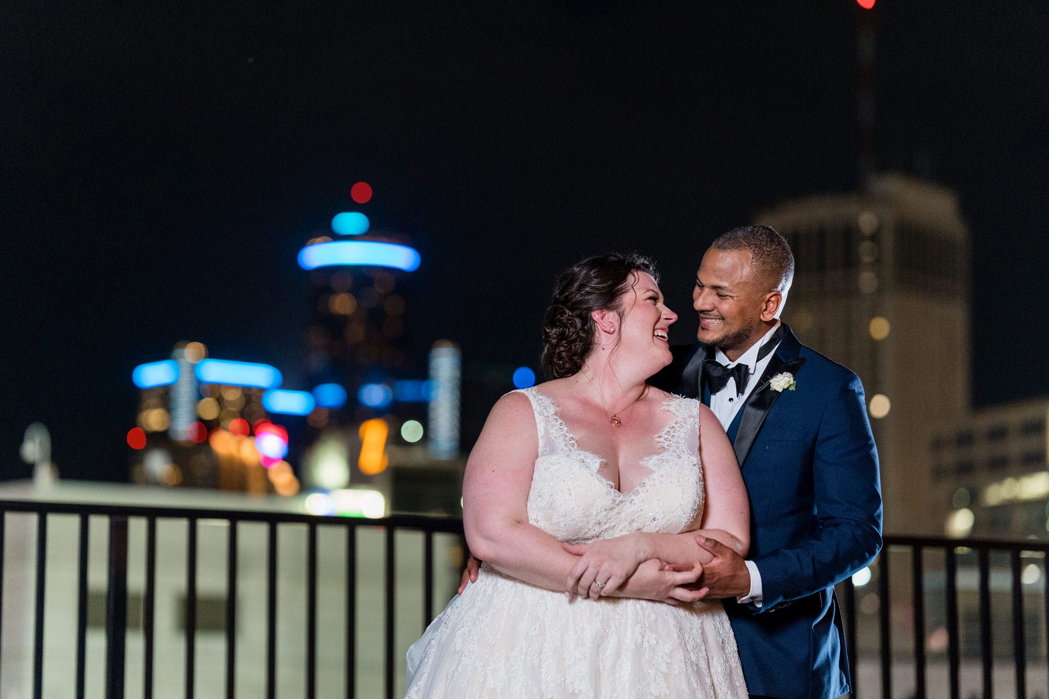 A nighttime photo of a groom hugging a bride from behind with the GM building in the background