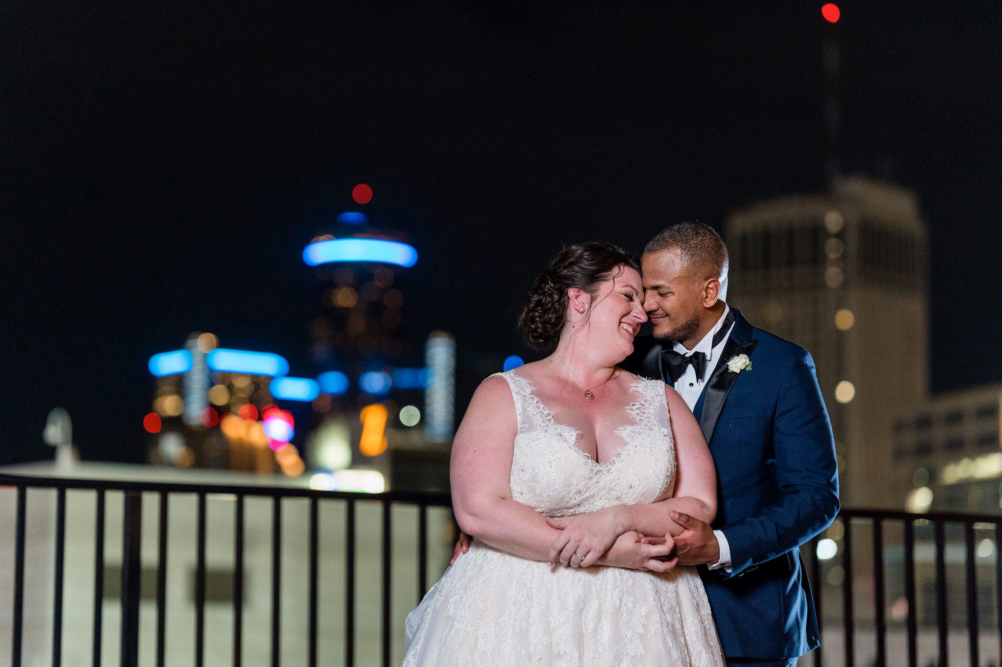 A nighttime photo of bride and groom snuggling up, nose to nose with the GM building in the background