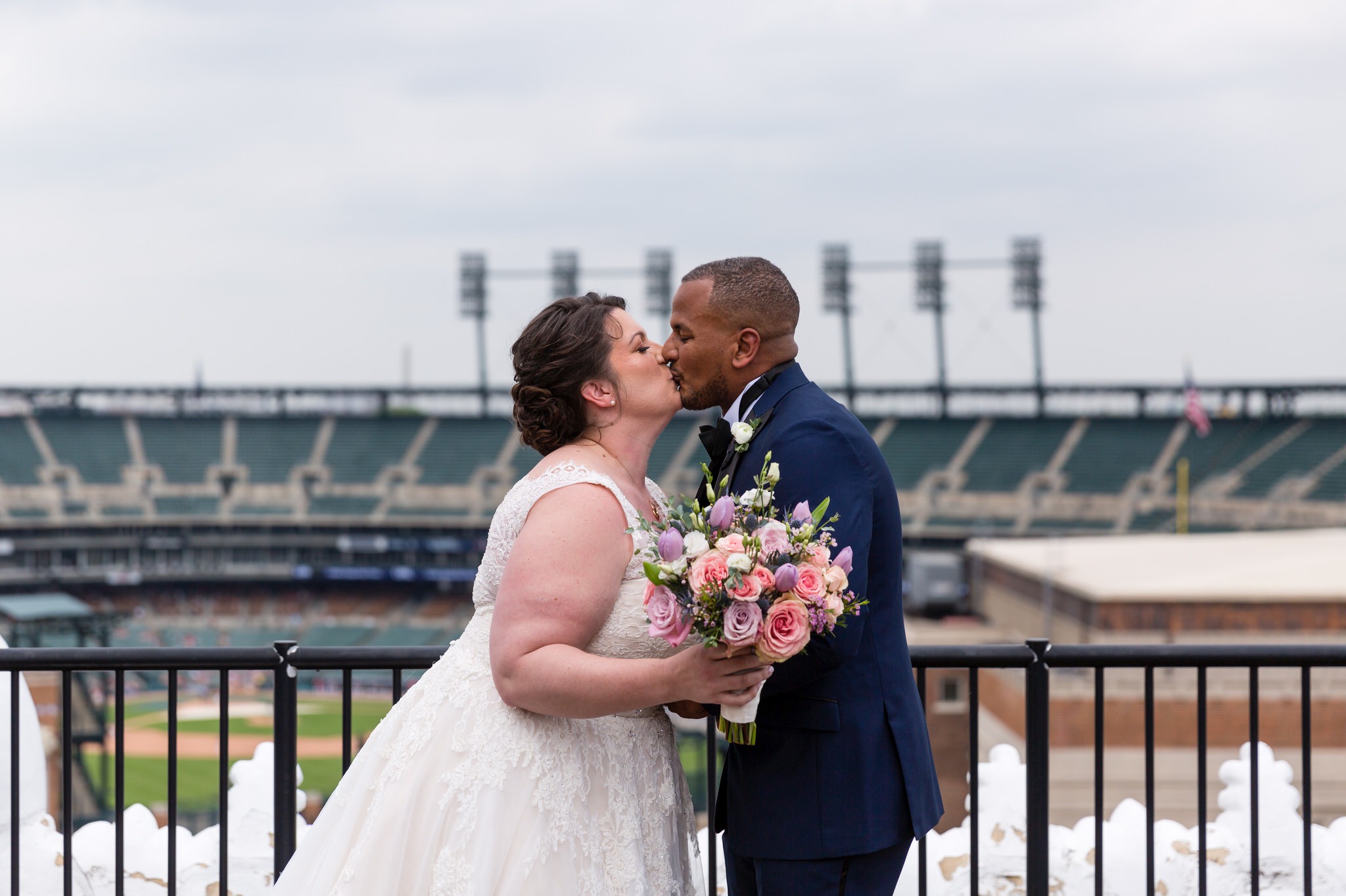 a bride and groom share a kiss  standing on top of the Detroit Opera house rooftop with Comerica Park in the background