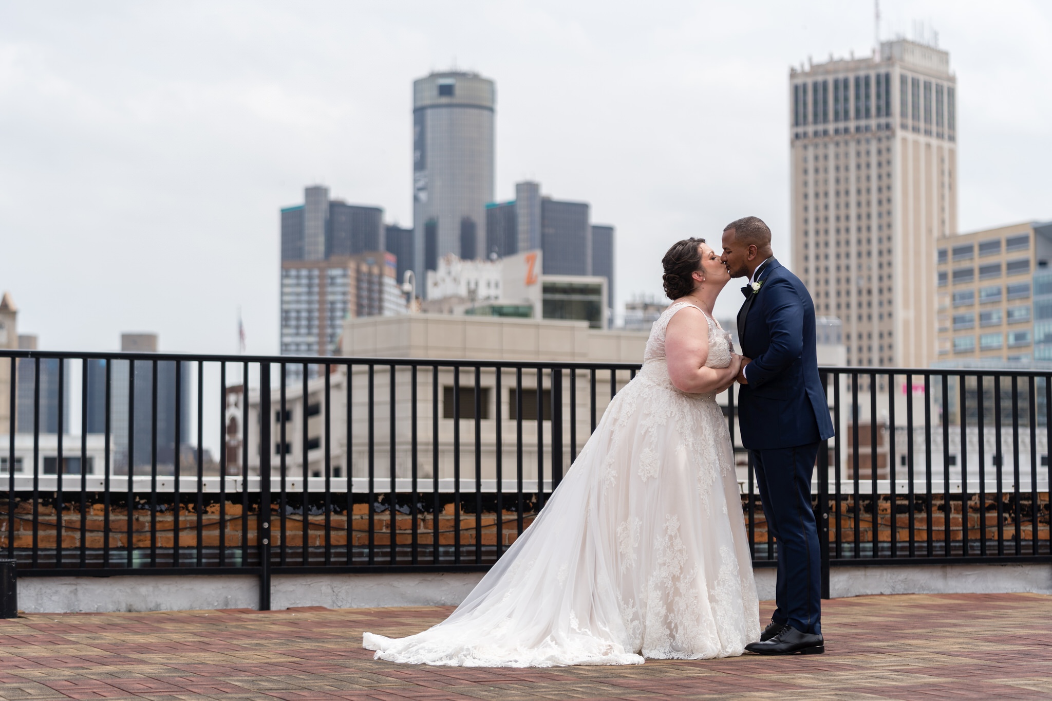 a bride and groom share a kiss  standing on top of the Detroit Opera house rooftop with the GM Headquarters in the background