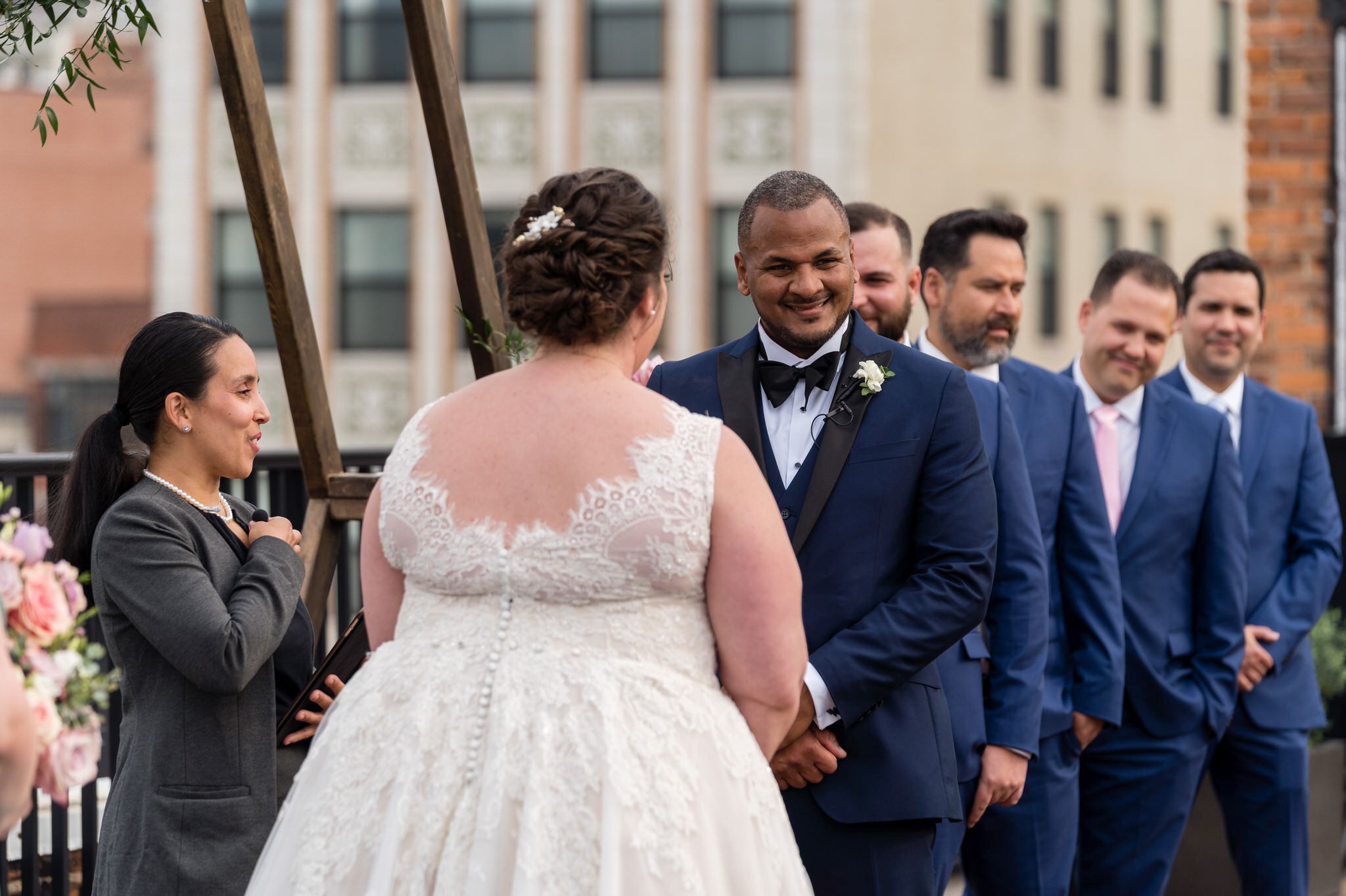 a groom, dressed in a blue tuxedo, smiles at his bride with groomsmen in the background