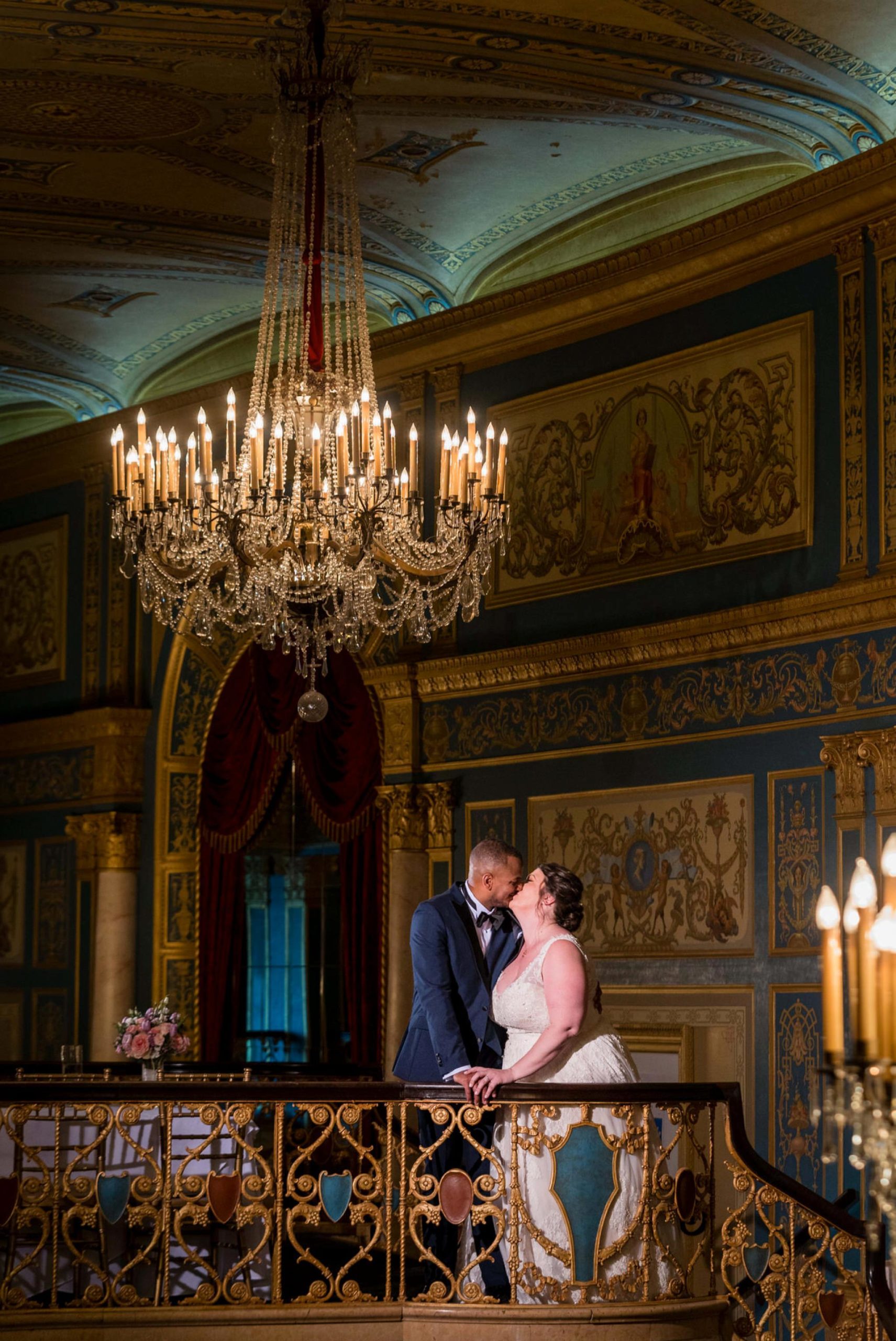 a bride and groom kiss holding an ornate railing with chandeliers in the foreground and background at the Detroit Opera House