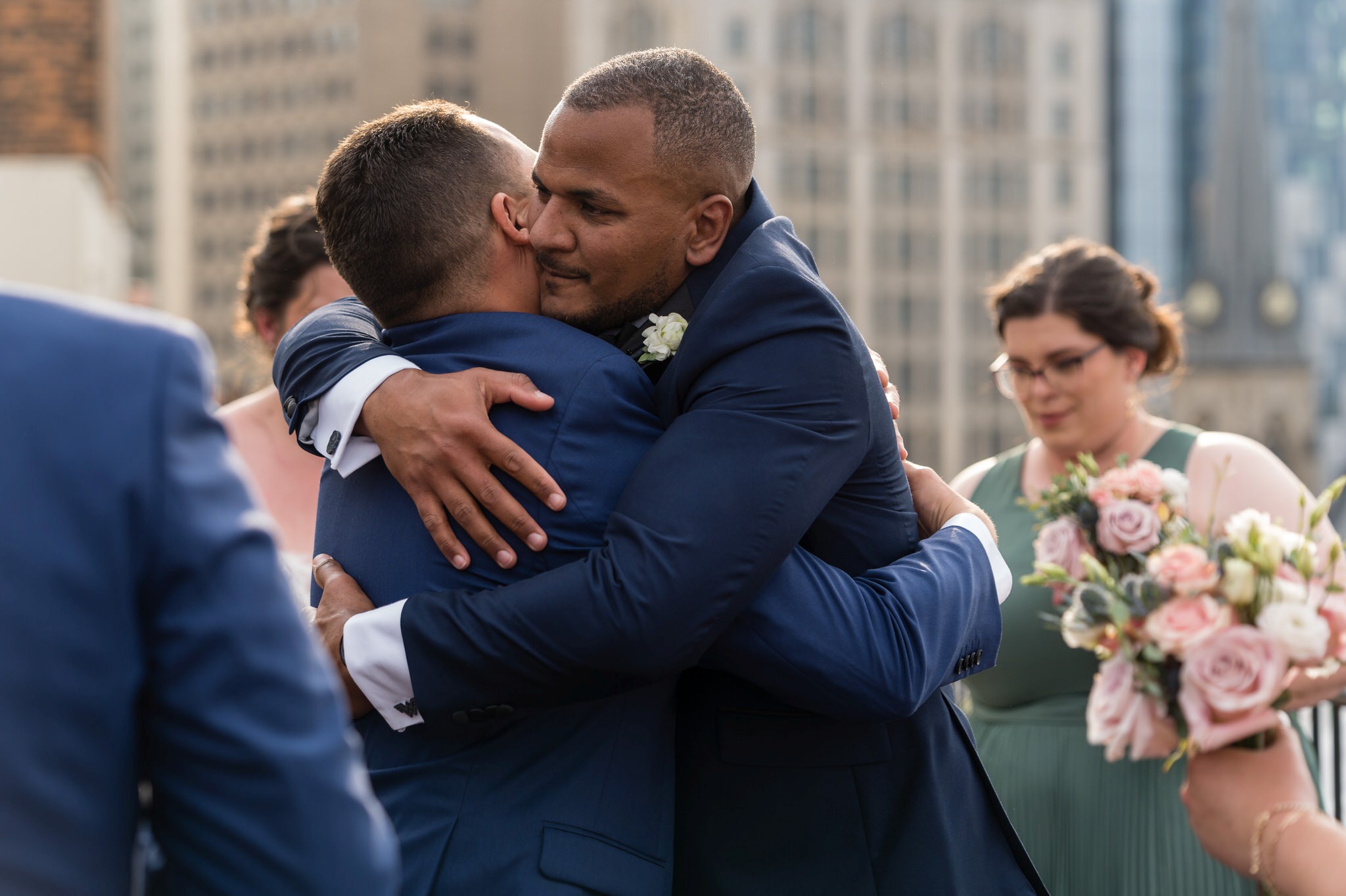 a groom, wearing a blue suit, hugs his groomsman after getting married at their rooftop wedding at the Detroit Opera House