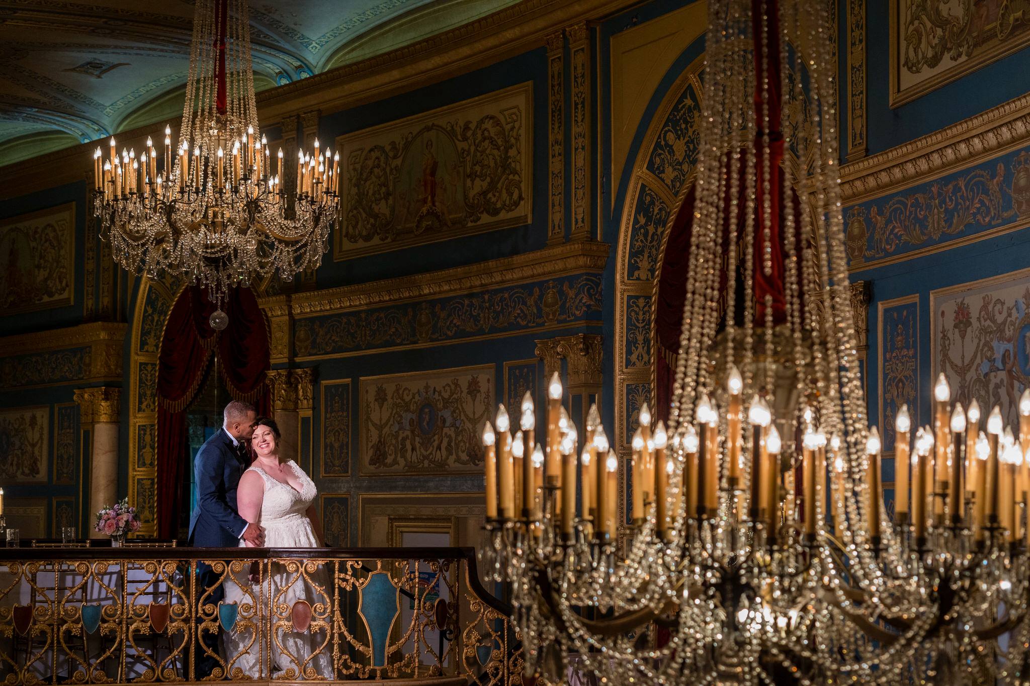 a bride and groom stand with chandeliers in the foreground and background at the Detroit Opera House
