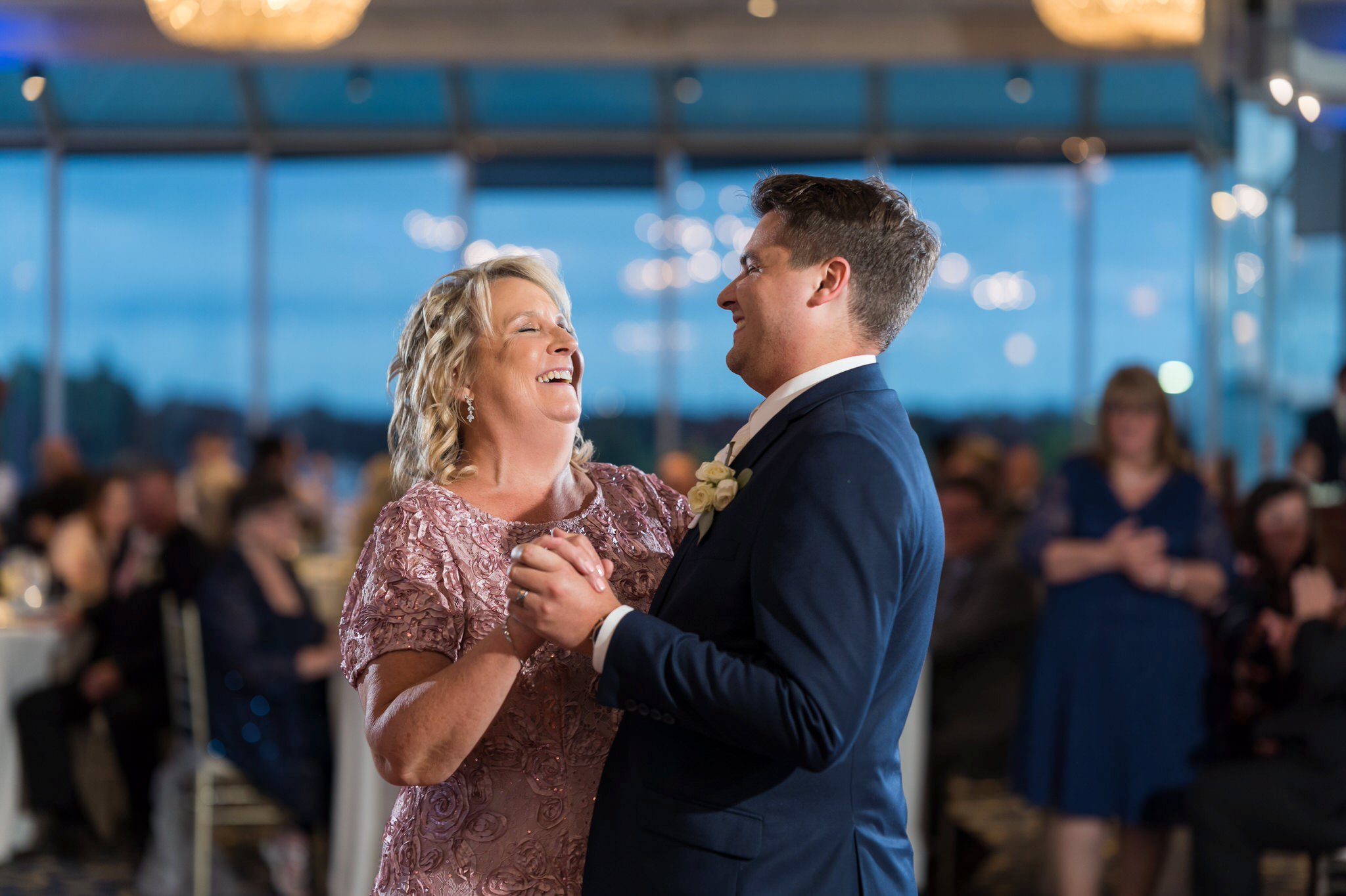 Mother of the groom, wearing a pink dress, dances with her son during MacRay Harbor wedding reception