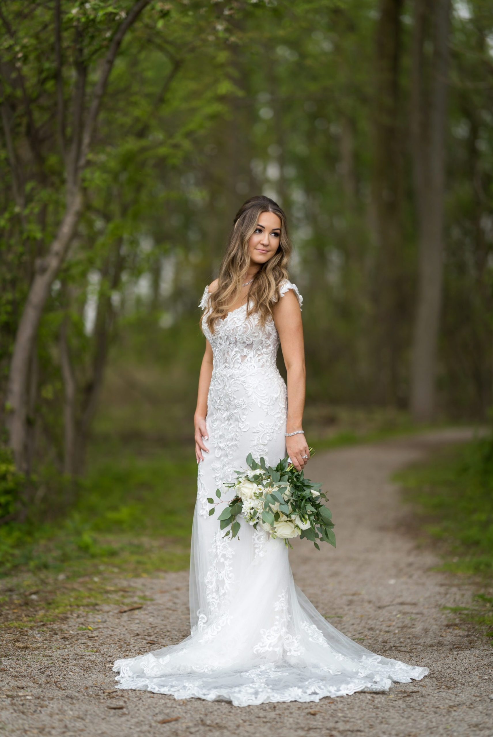a bride poses, looking over her shoulder, in nature while holding her white rose bouquet