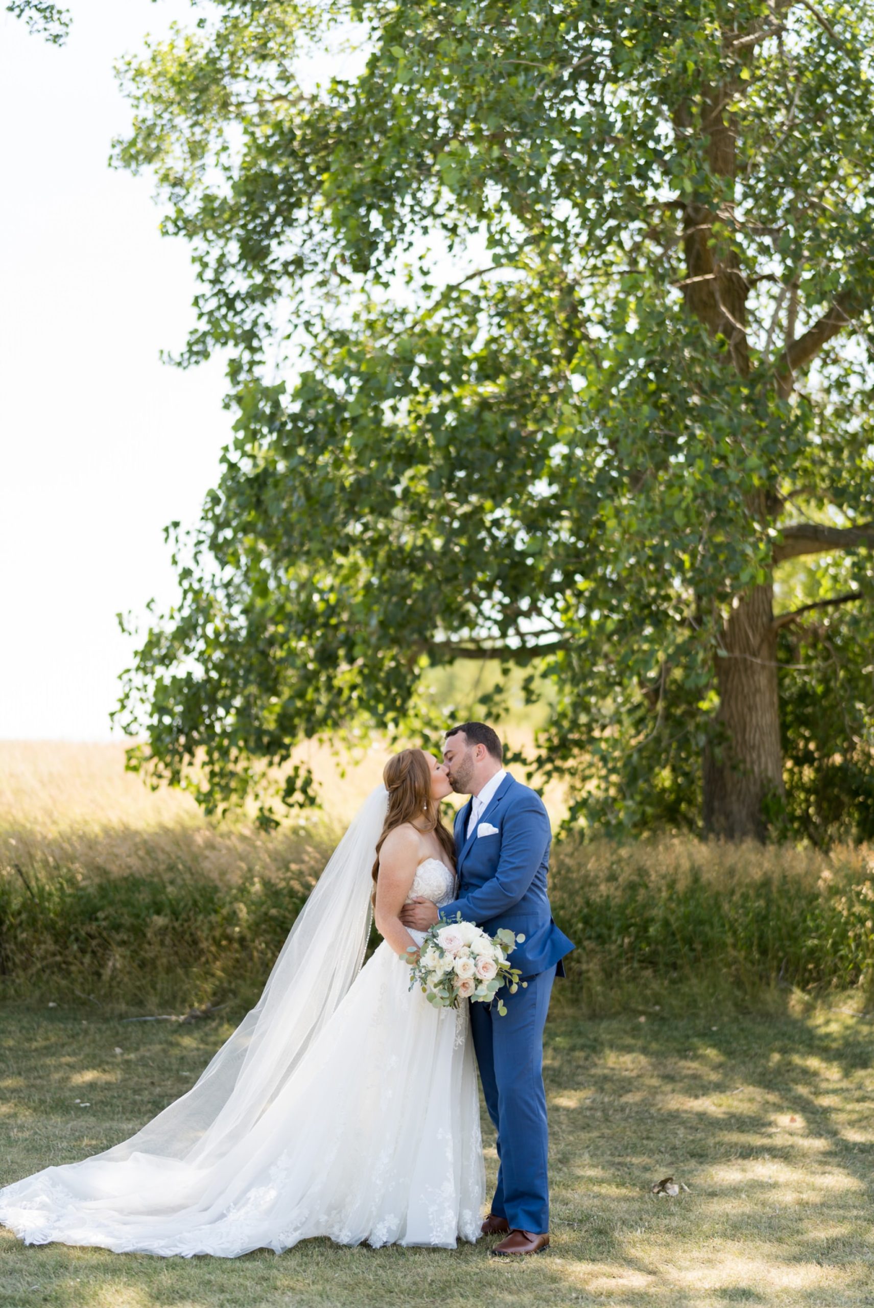 Sarah and Justin kiss near a field of grass and a tree on their wedding day. 