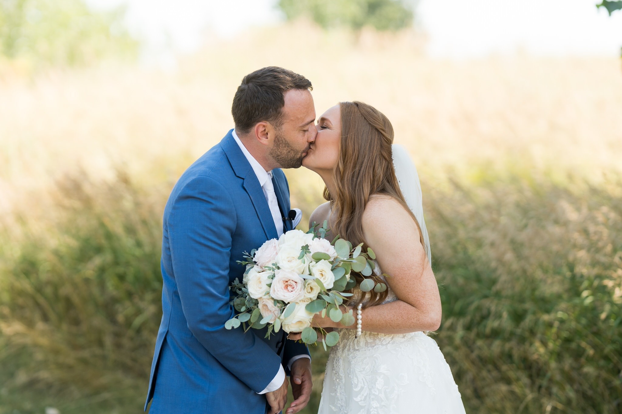 Sarah and Justin kiss on their wedding day, standing in front of a field of wild grass. 