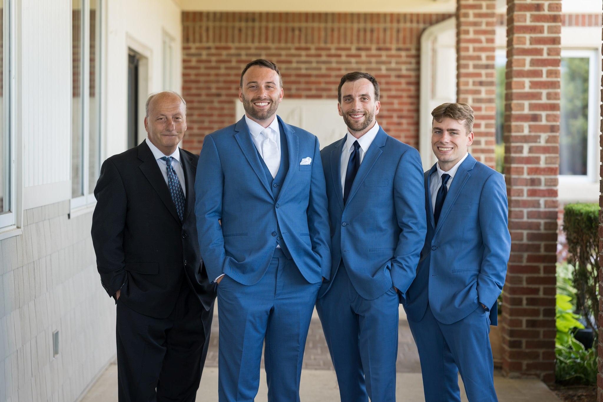 A groom wearing a blue suit is flanked by his dad and brothers at his Greystone Golf Club wedding