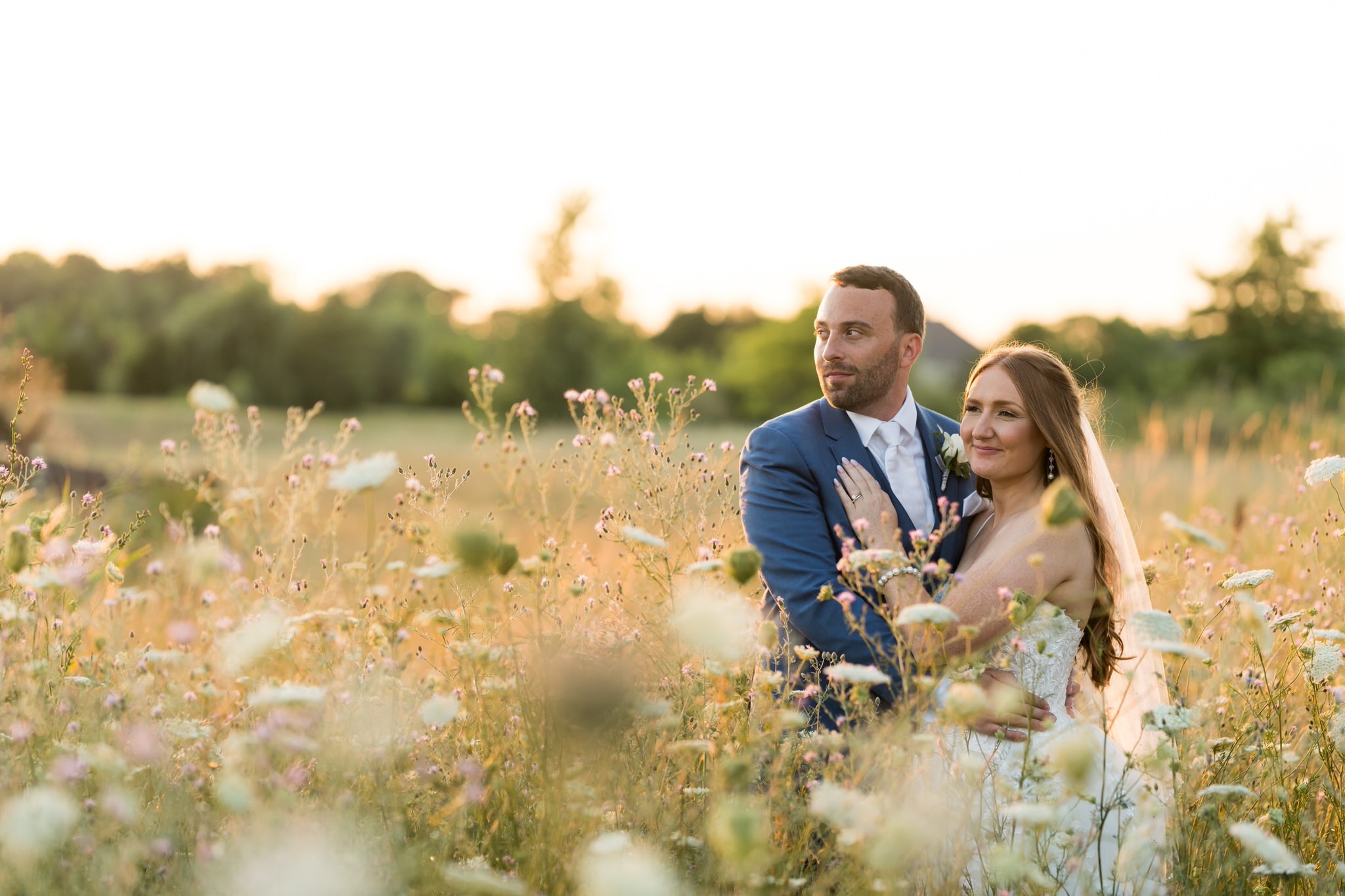 A bride and groom look outward in a field of wildflowers and tall grass during golden hour by Brian Weitzel Photography. 