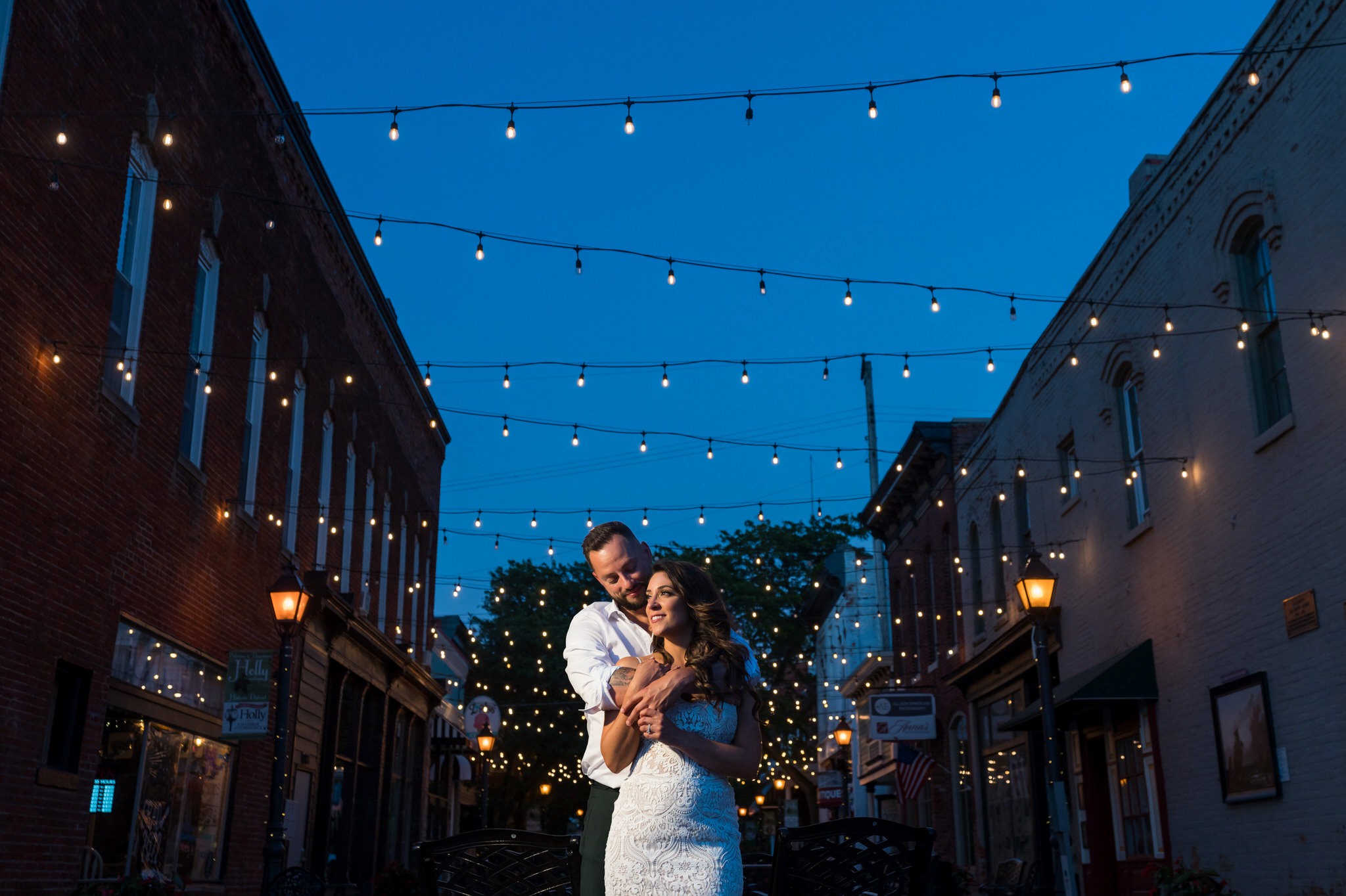 groom hugs the bride from behind under twinkle lights in downtown Holly at night