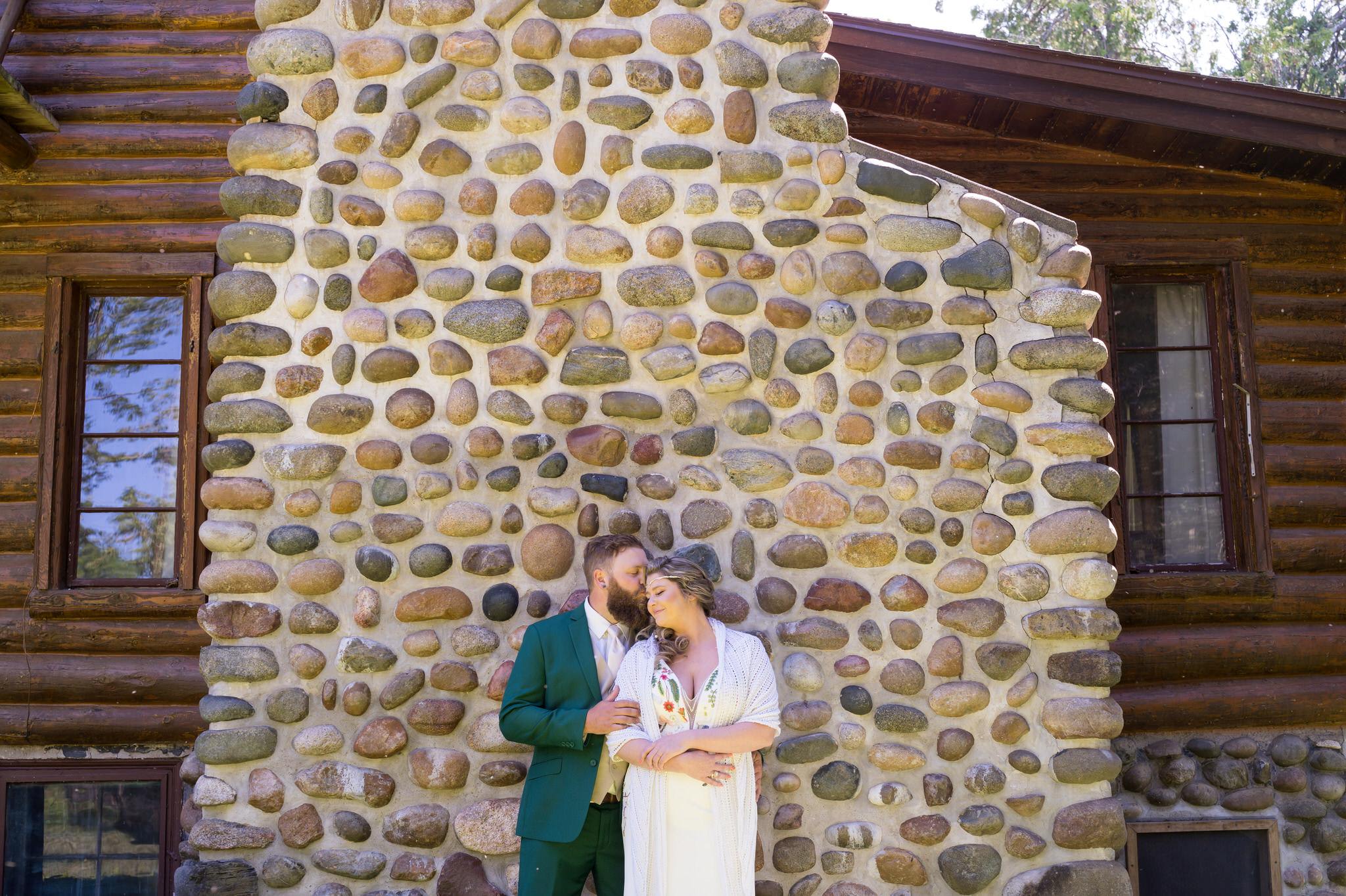 groom kisses bride on the head with  a stone chimney behind them
