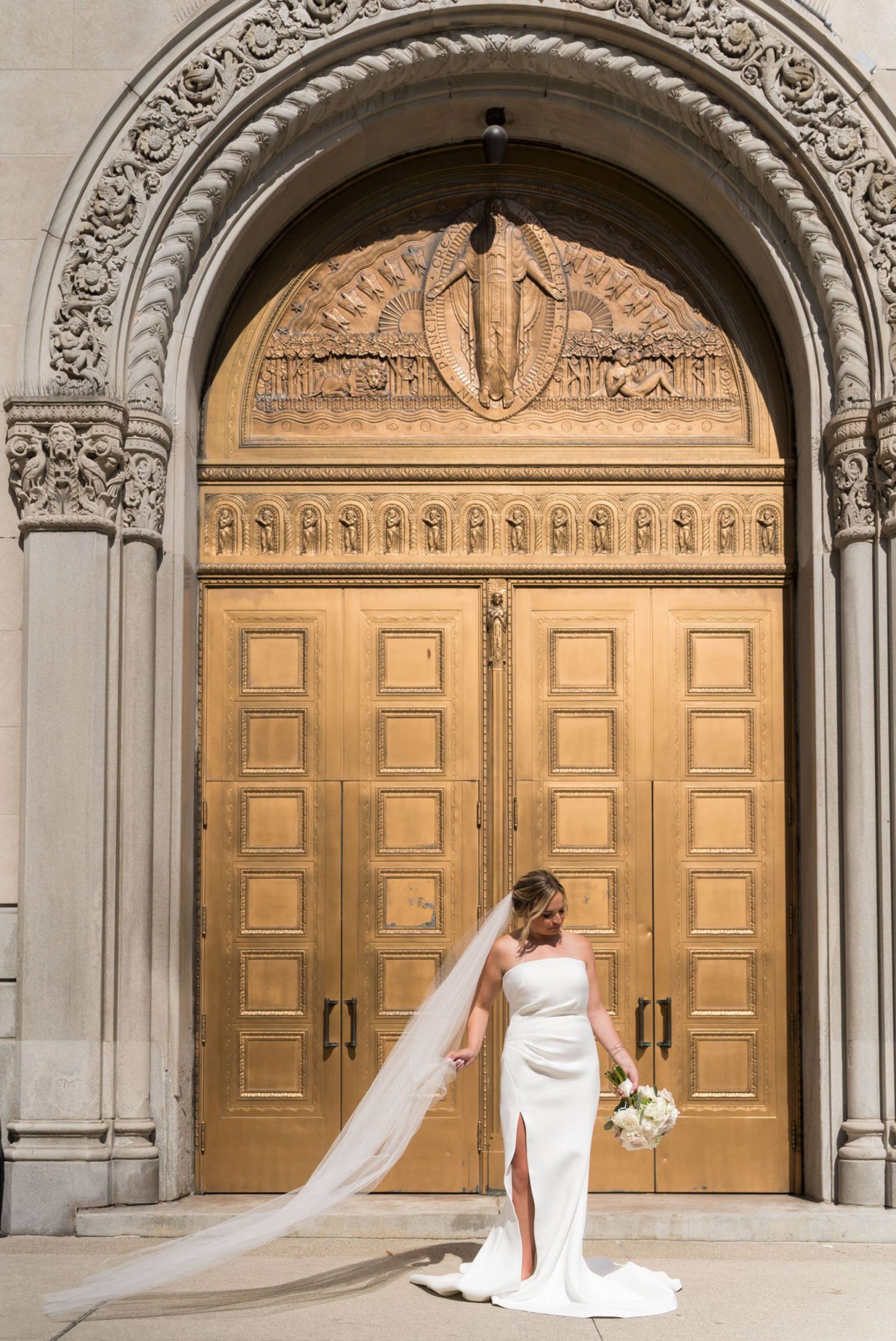 bride holds her veil as it flows in the wind in front of a gold door at St. Aloysius church in detroit
