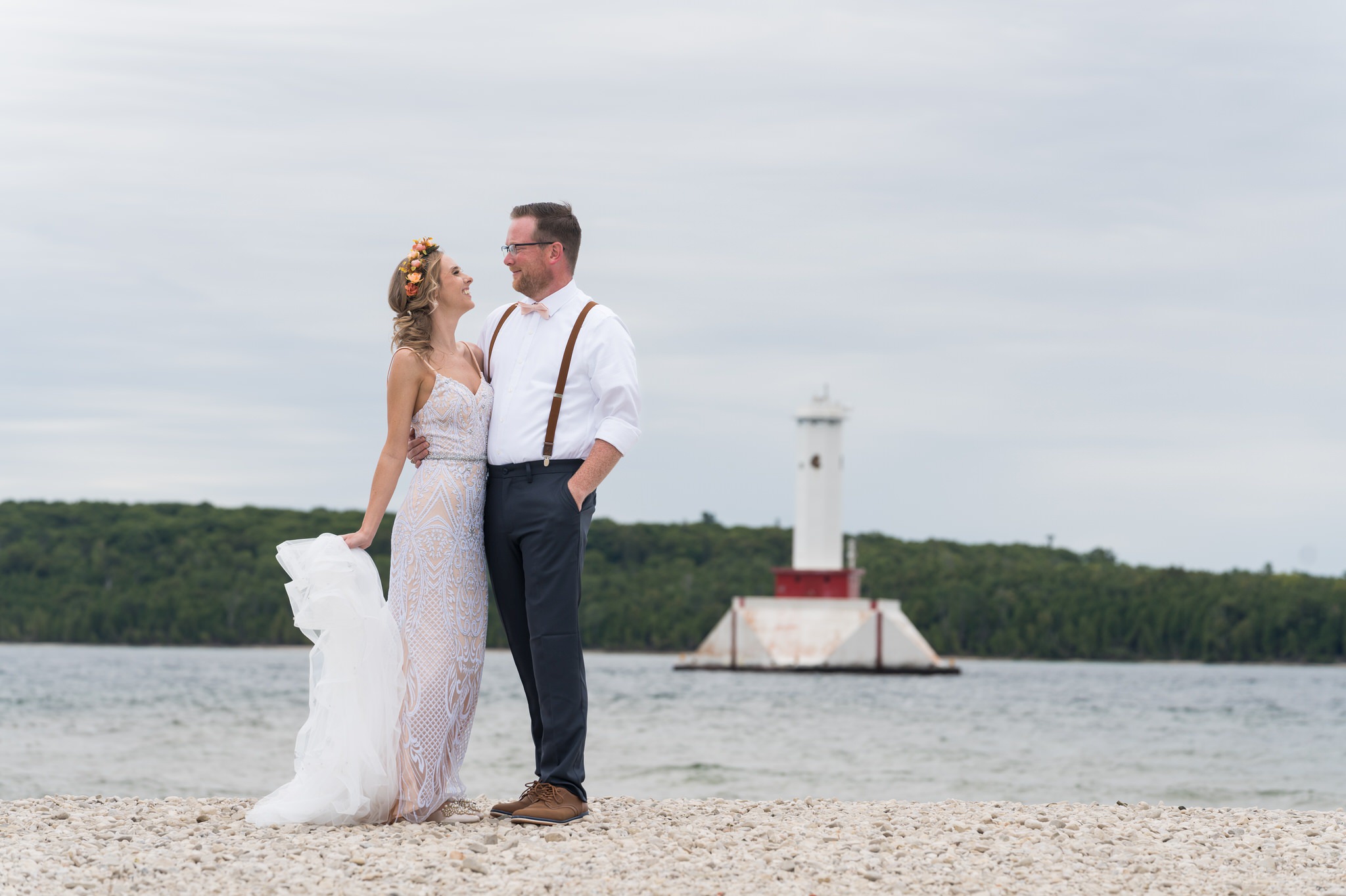 A bride and groom look at each other on the shores of Mackinac Island with a lighthouse in the background during their Mission Point Resort wedding.  