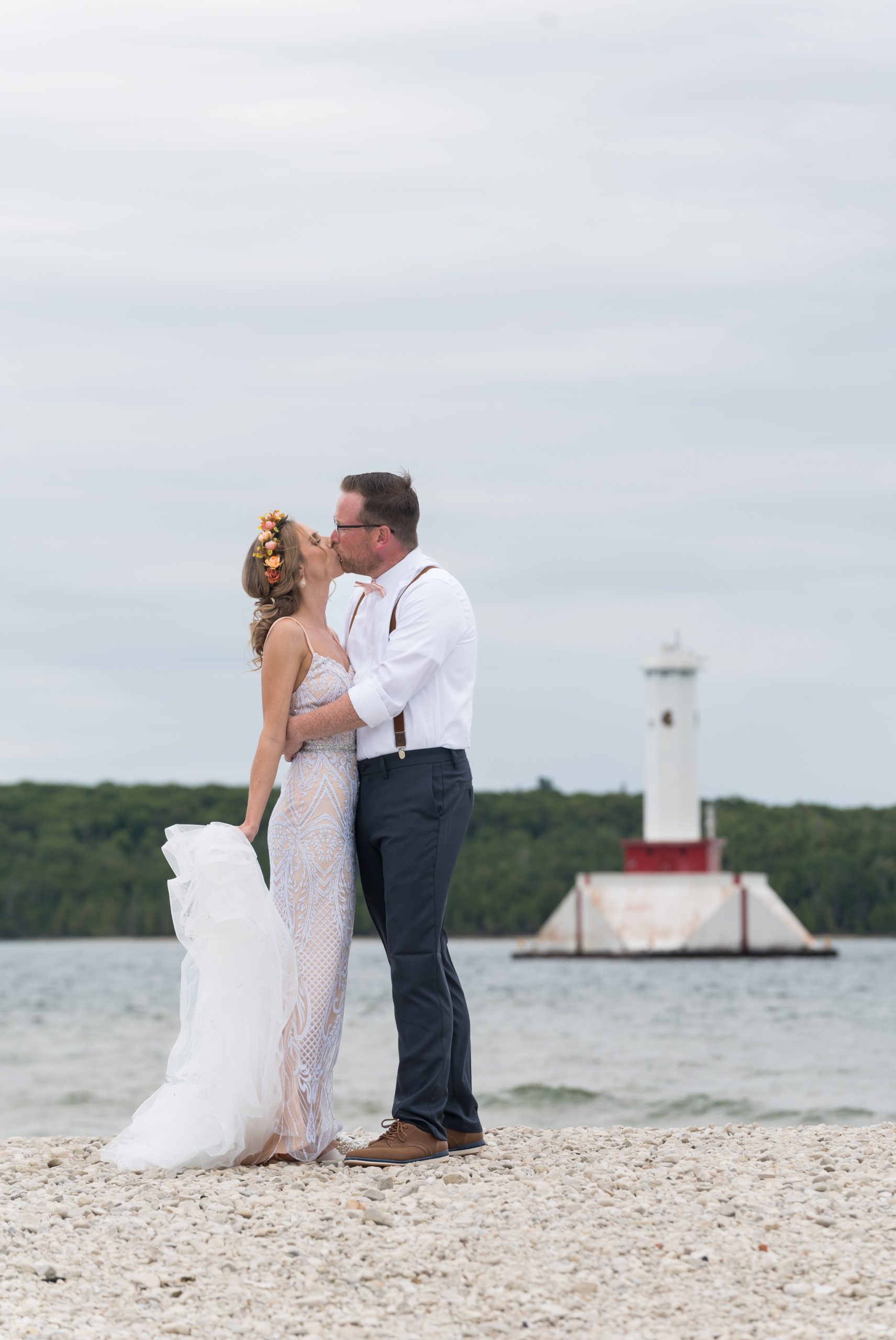 A bride and groom kiss on the shores of Mackinac Island with a lighthouse in the background during their Mission Point Resort wedding.  