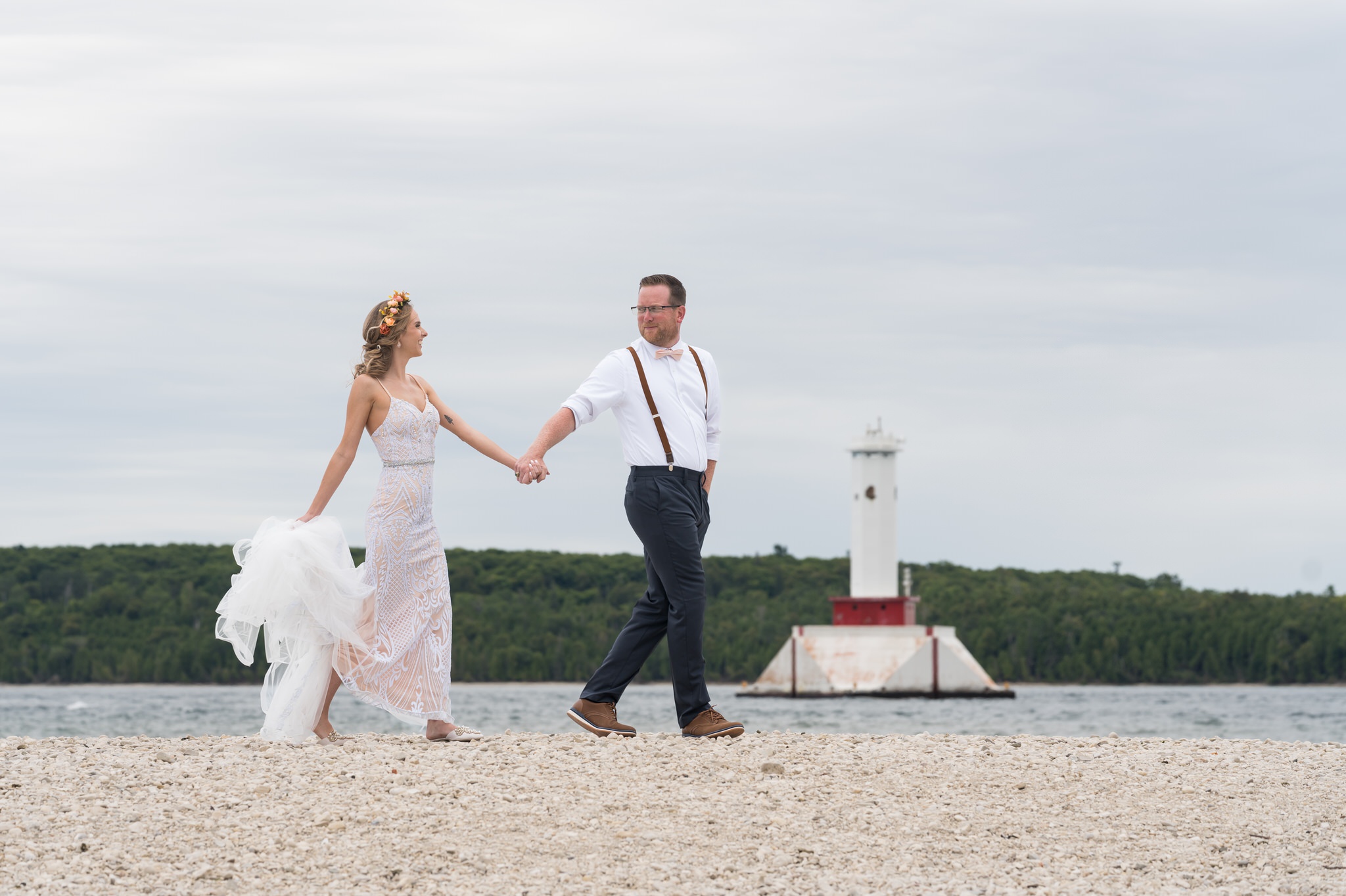 A bride and groom walk on the shores of Mackinac Island with a lighthouse in the background during their Mission Point Resort wedding.  