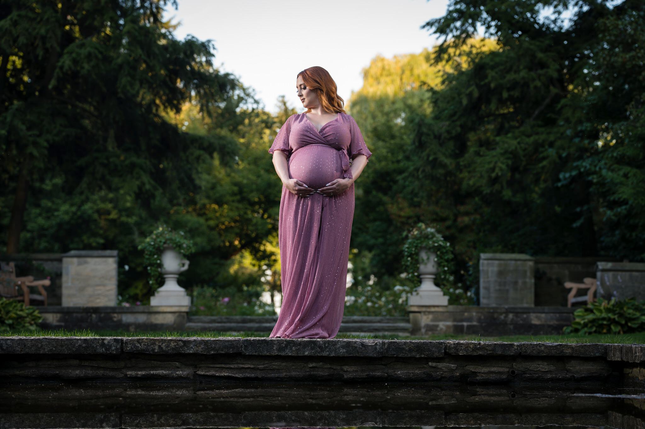 An expecting mom looks over her shoulder and holds her bump as her reflection is seen in a pond during her maternity session at Edsel Ford