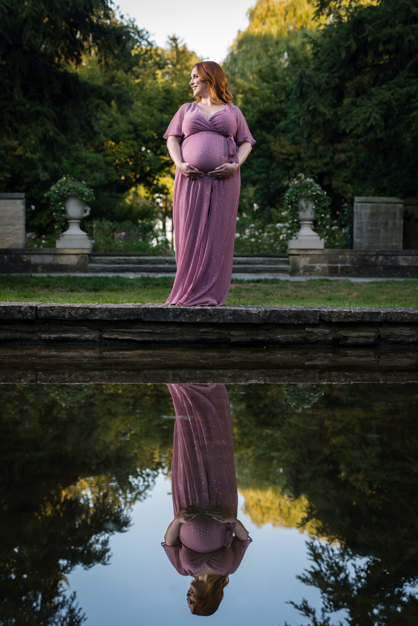 An expecting mom, wearing a purple dress holds her bump as her reflection is seen in  a pond during her maternity session at Edsel Ford