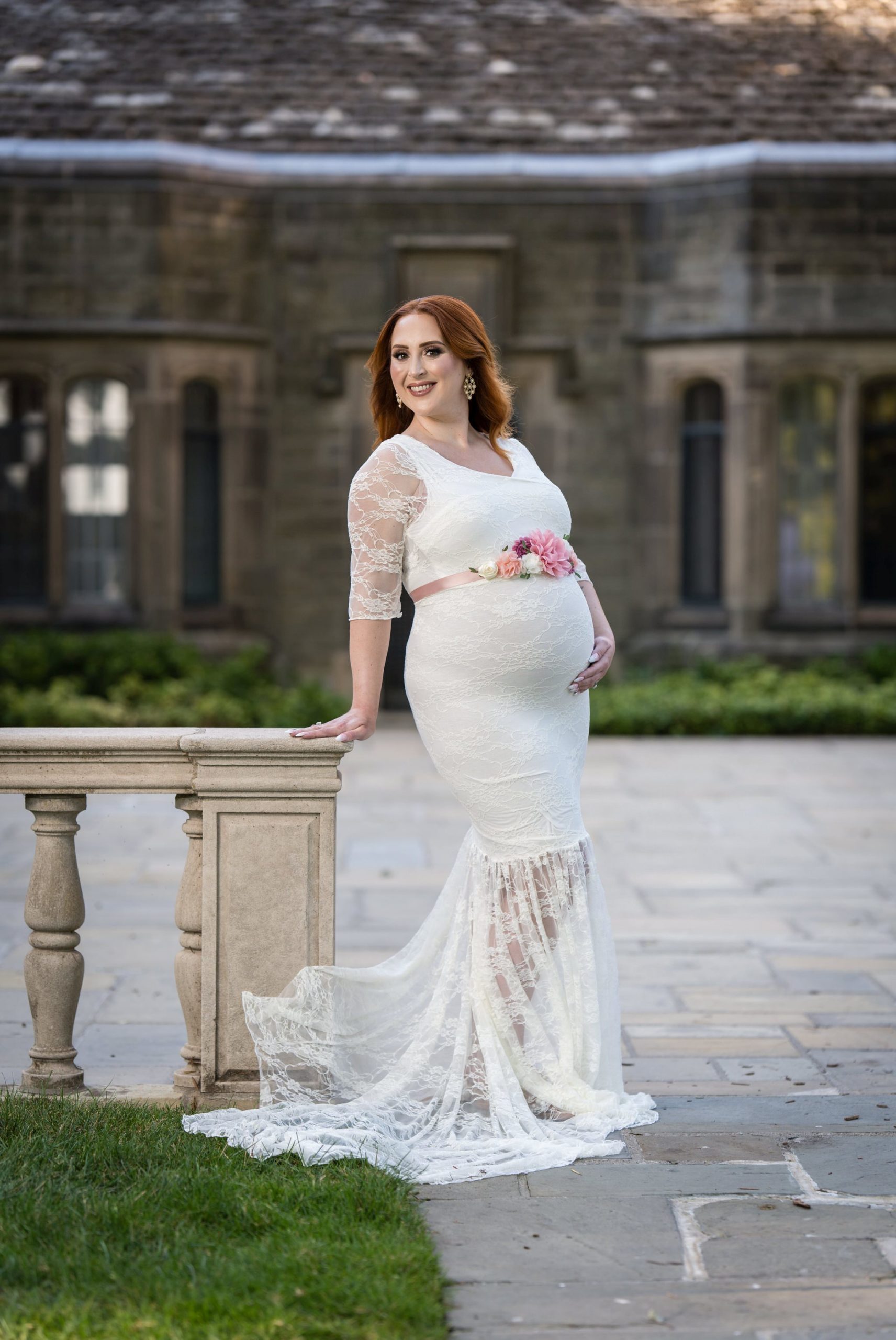 A pregnant woman wearing a white lace dress holds her bump during a maternity session at Edsel Ford House.