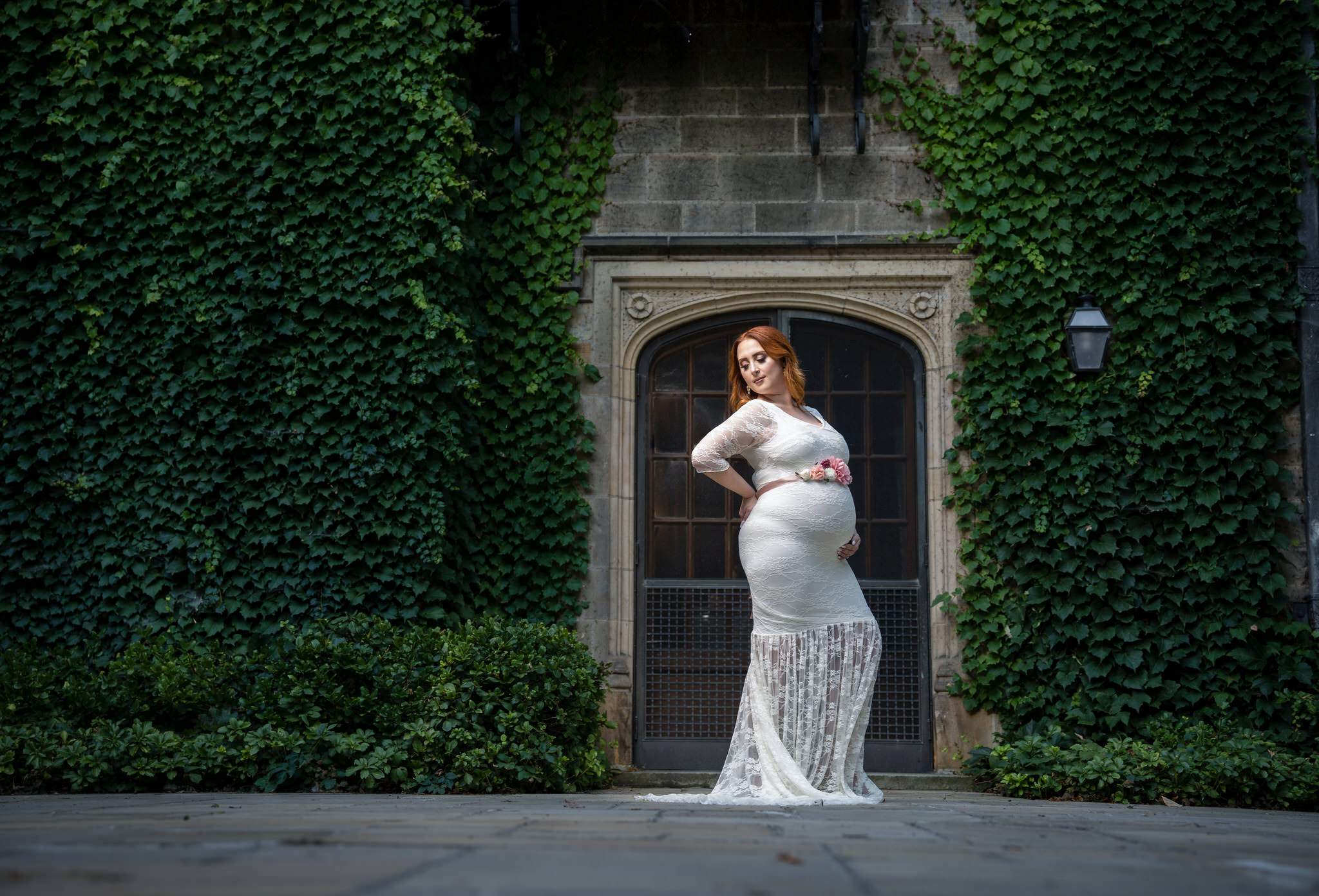 An expecting mom holds her stomach wearing a white flowing dress at the Edsel Ford House