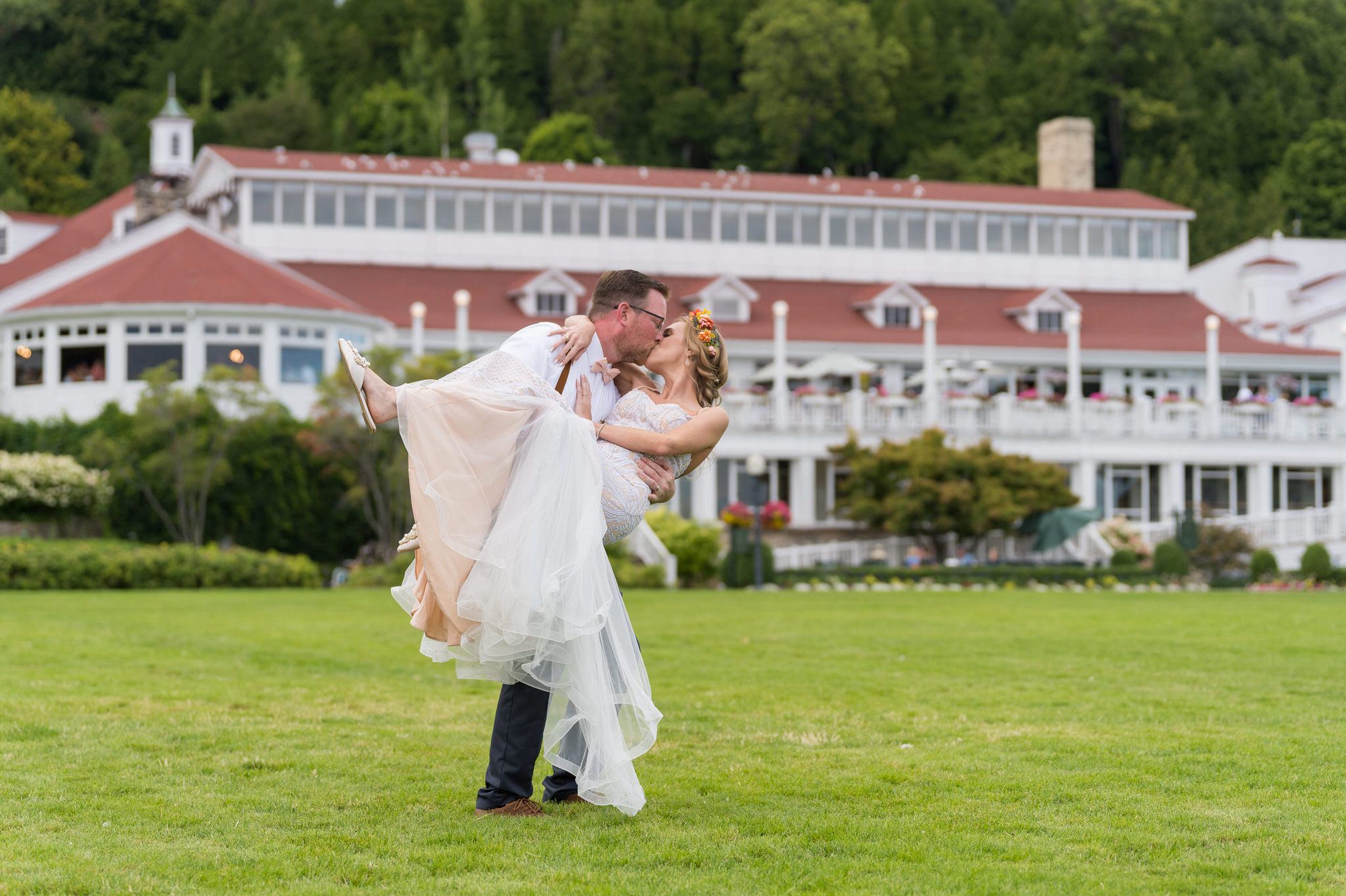 A groom, carrying his bride, kisses her with their Mission Pointe Resort wedding in the background.  