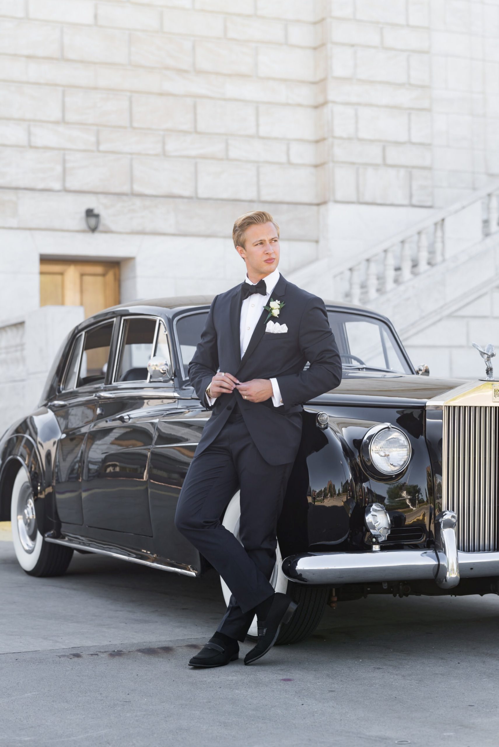 A groom leans against a black Rolls Royce while buttoning his tuxedo jacket.    