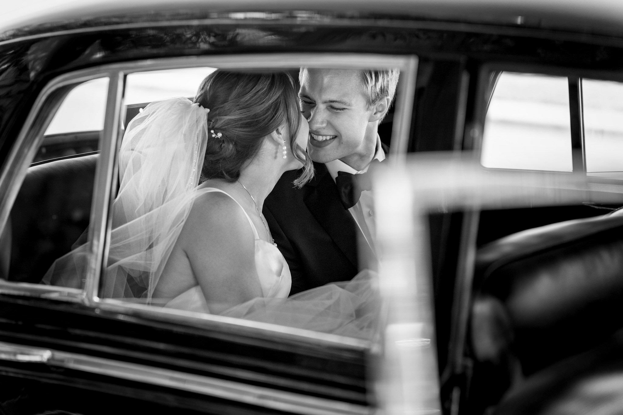 Shot through the car window, a groom smiles at his bride on their wedding day.  