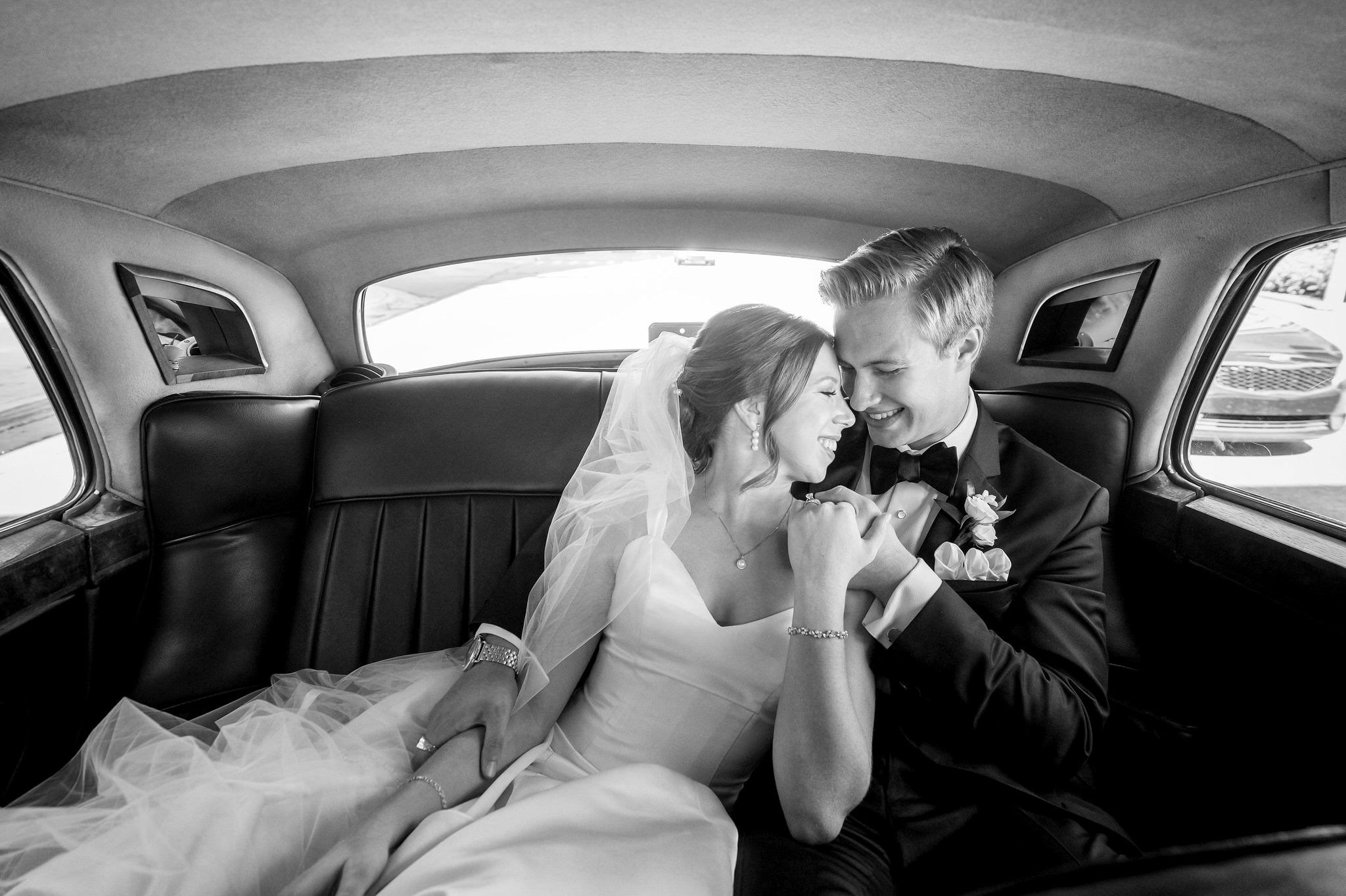 A couple smile and snuggle while sitting in the back of a Rolls Royce on their wedding day.  