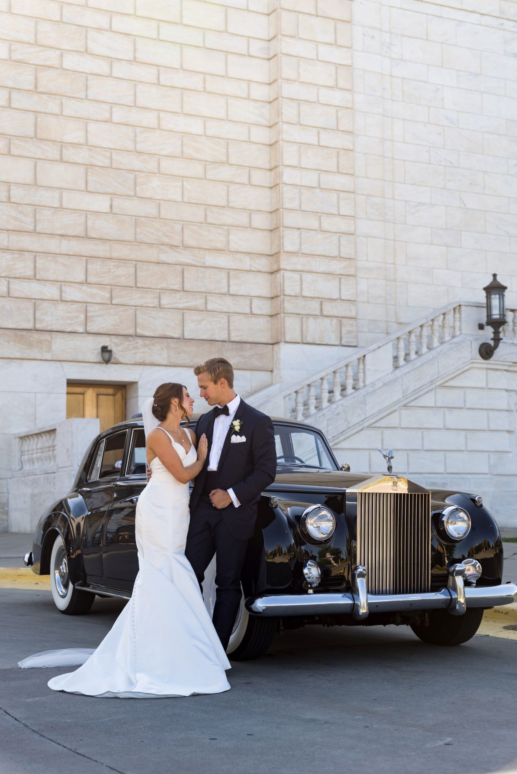 A groom relaxes against a black Rolls Royce with one hand in his pocket.  