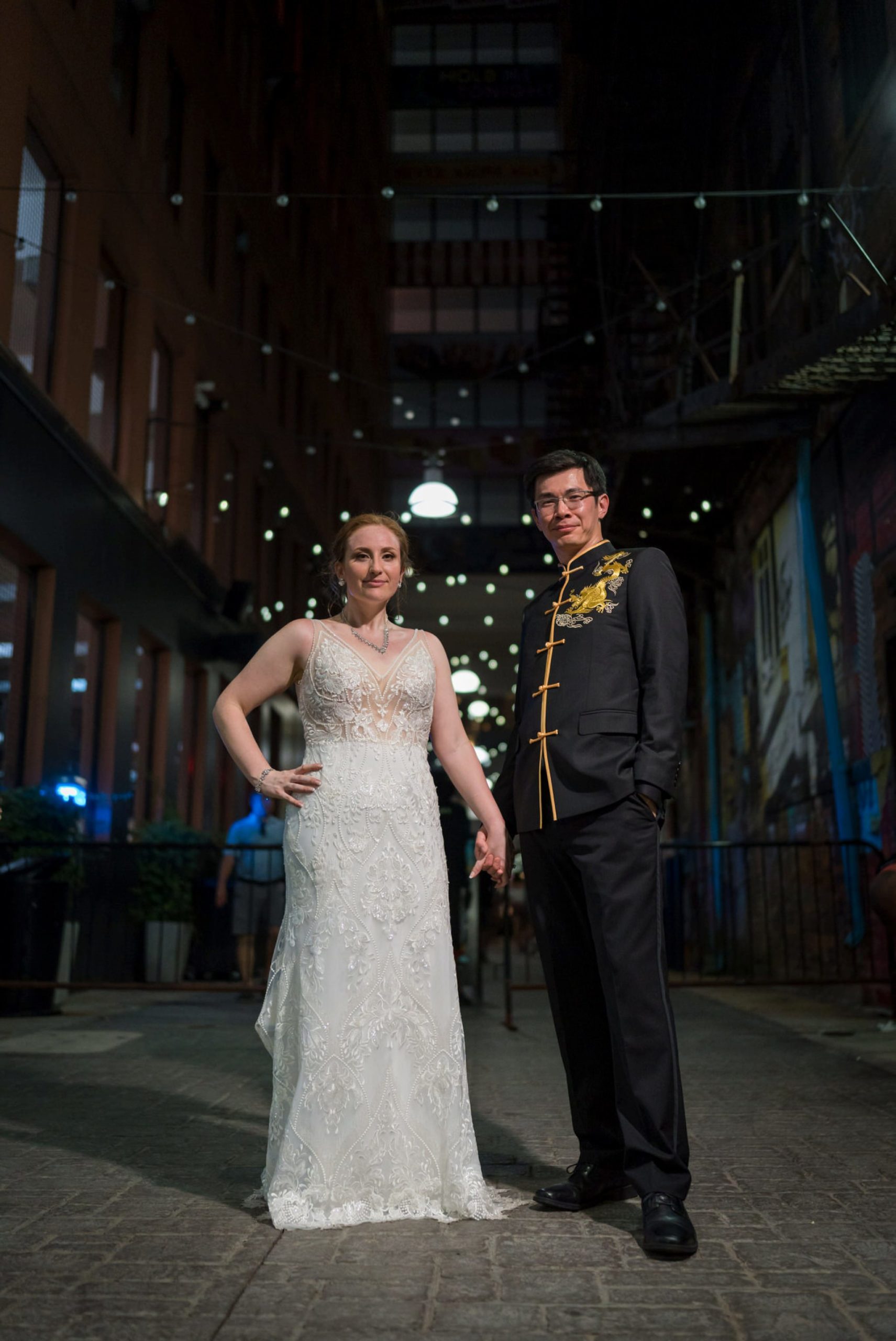 bride and groom, wearing traditional Chinese wedding garments, hold hands at night in Detroit's Z lot. 