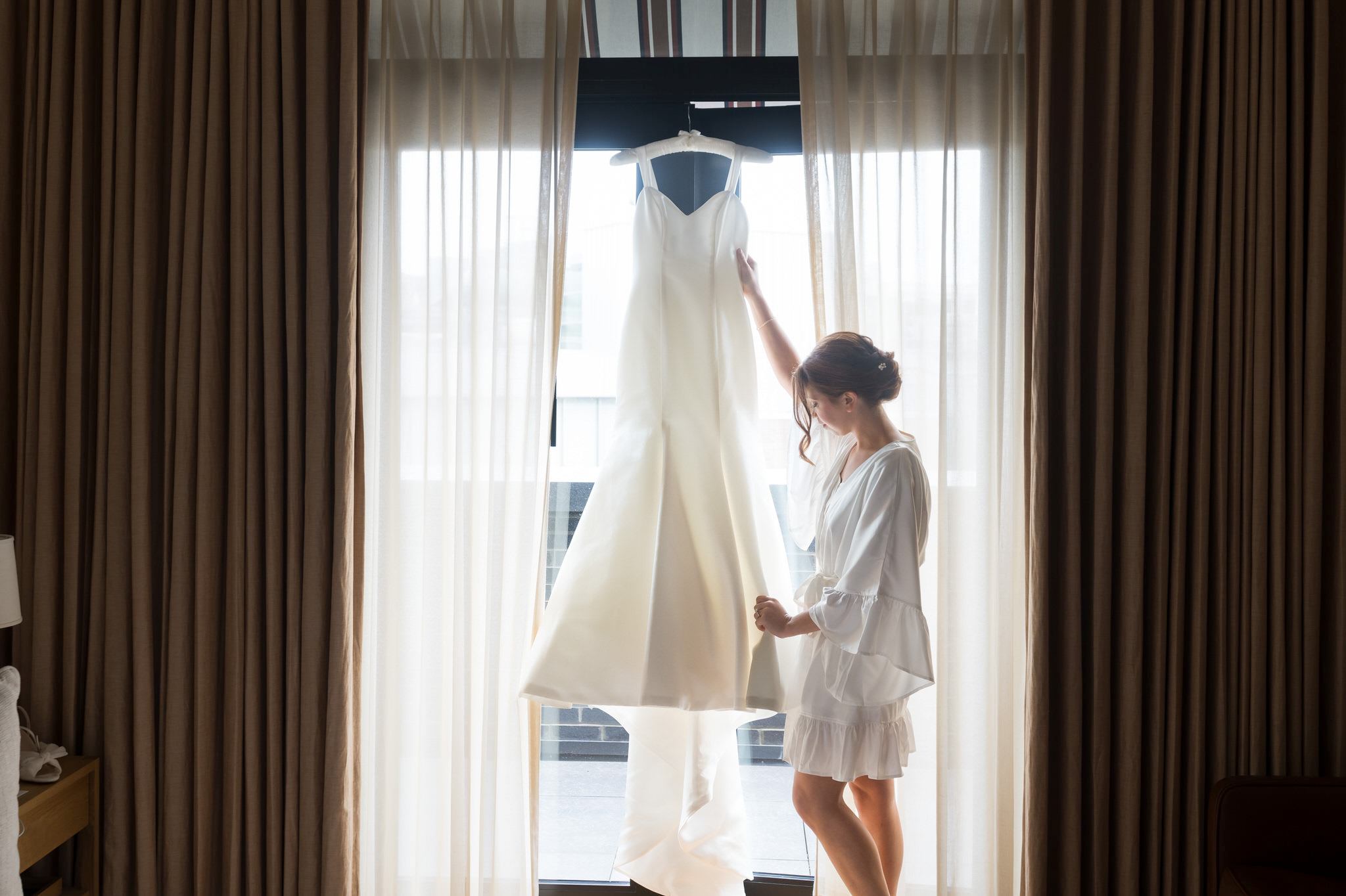 A bride wearing a white silk robe examines her hanging wedding dress in a Shinola Hotel room.  