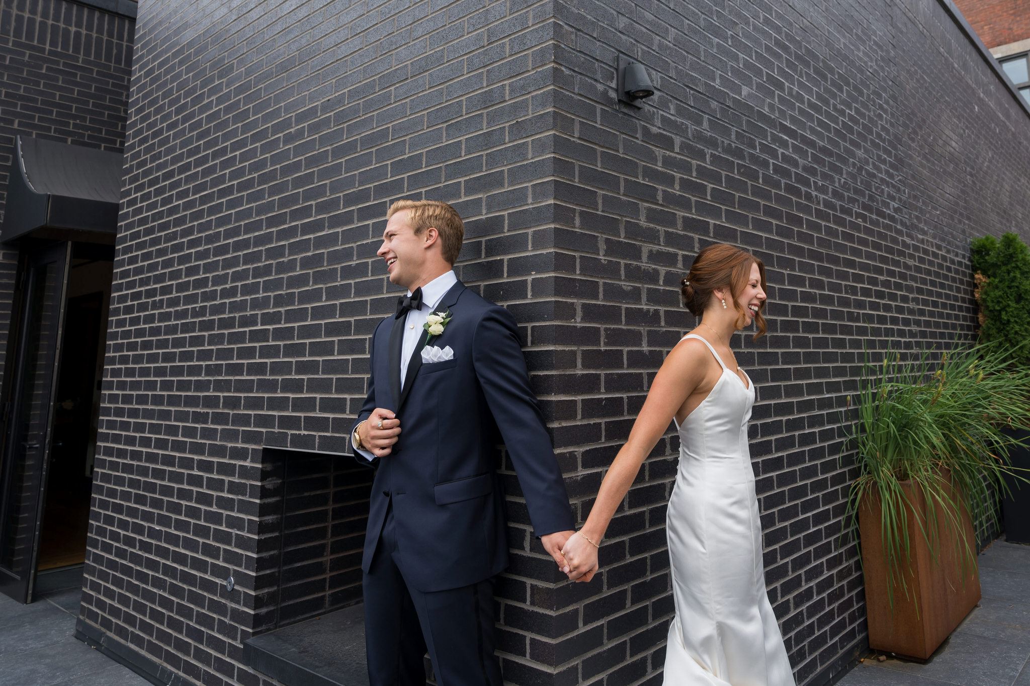 A bride and groom hold hands while hiding behind a corner on their wedding day.  