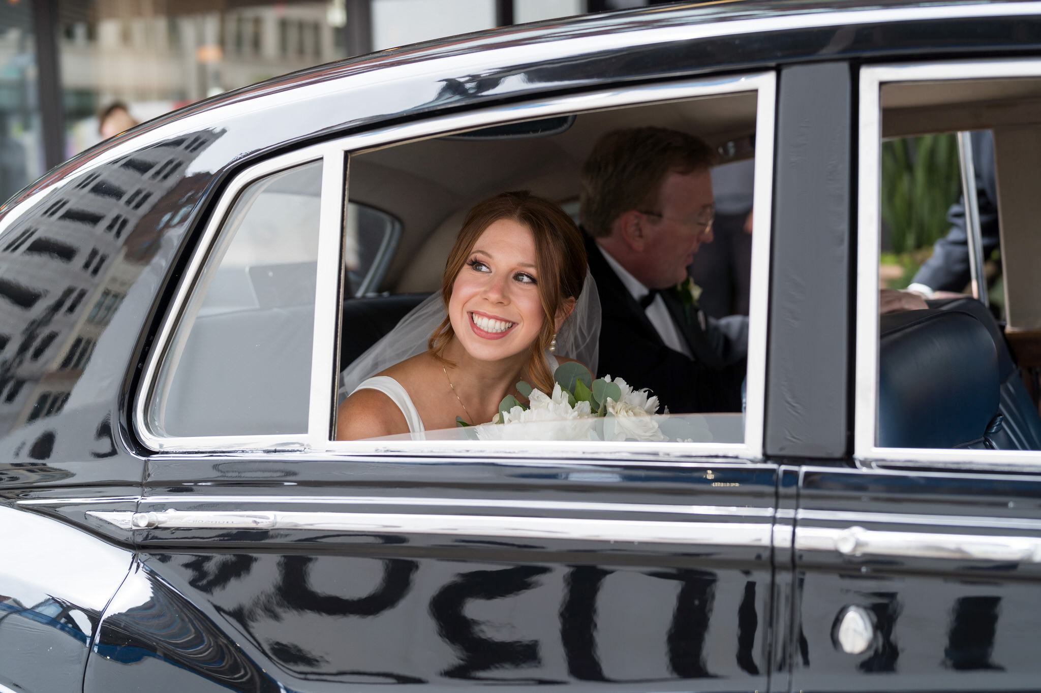 A red-headed bride smiles looking out of the window of a 1958 black Rolls Royce on her wedding day.  