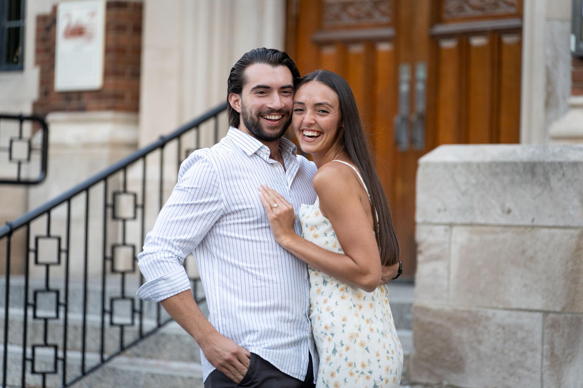 a couple smile and laugh, embracing each other and facing the camera during a downtown Birmingham proposal 