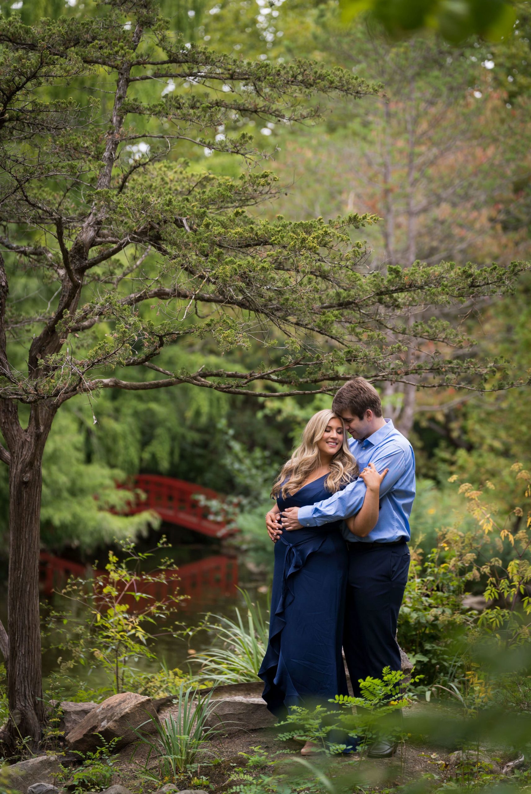 A couple hug with a red bridge in the background during their Cranbrook engagement session.  