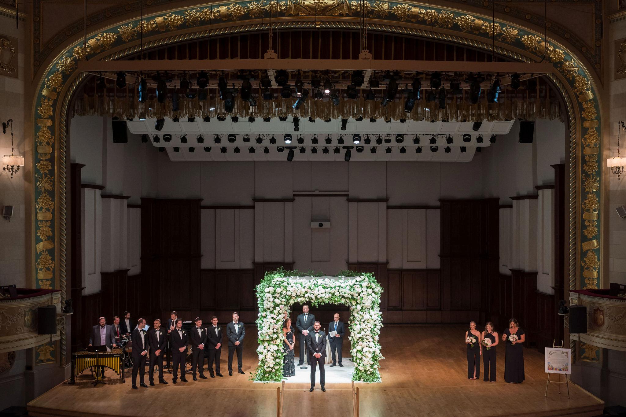 A groom waits, standing in front of a white floral chuppah at his Detroit Orchestra Hall wedding.  