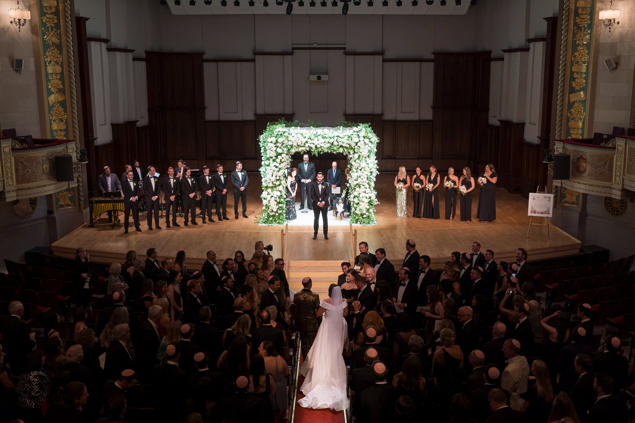 A bride is escorted to her groom, who waits near a white floral chuppah on stage at a Detroit Orchestra Hall wedding.  