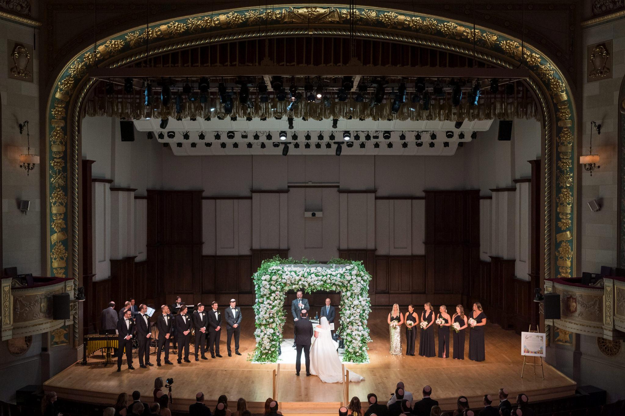 A bride and groom stand beneath an floral chuppah on stage at a Detroit Orchestra Hall wedding.  