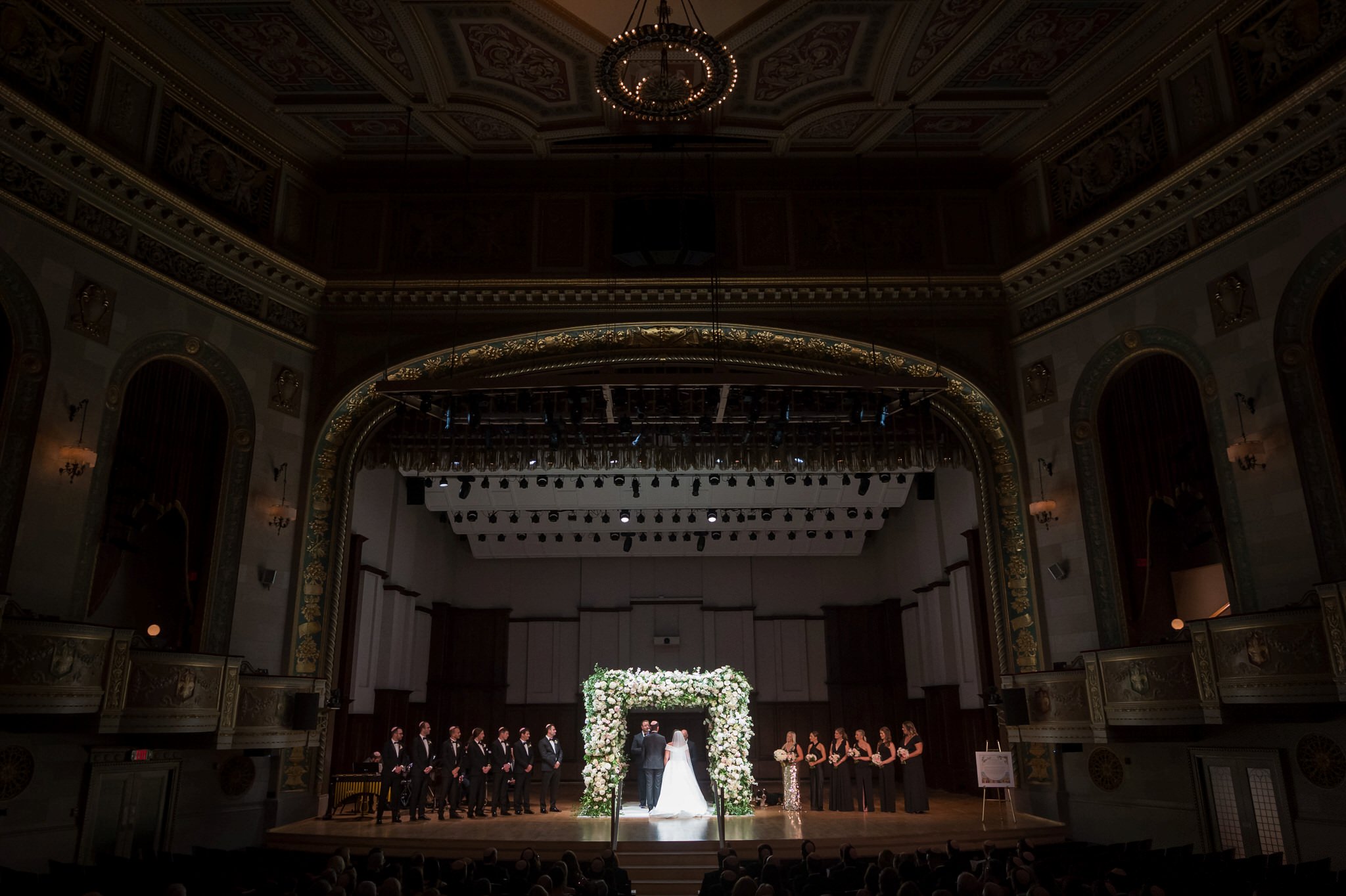 A bride and groom stand beneath an floral chuppah on stage at a Detroit Orchestra Hall wedding.  