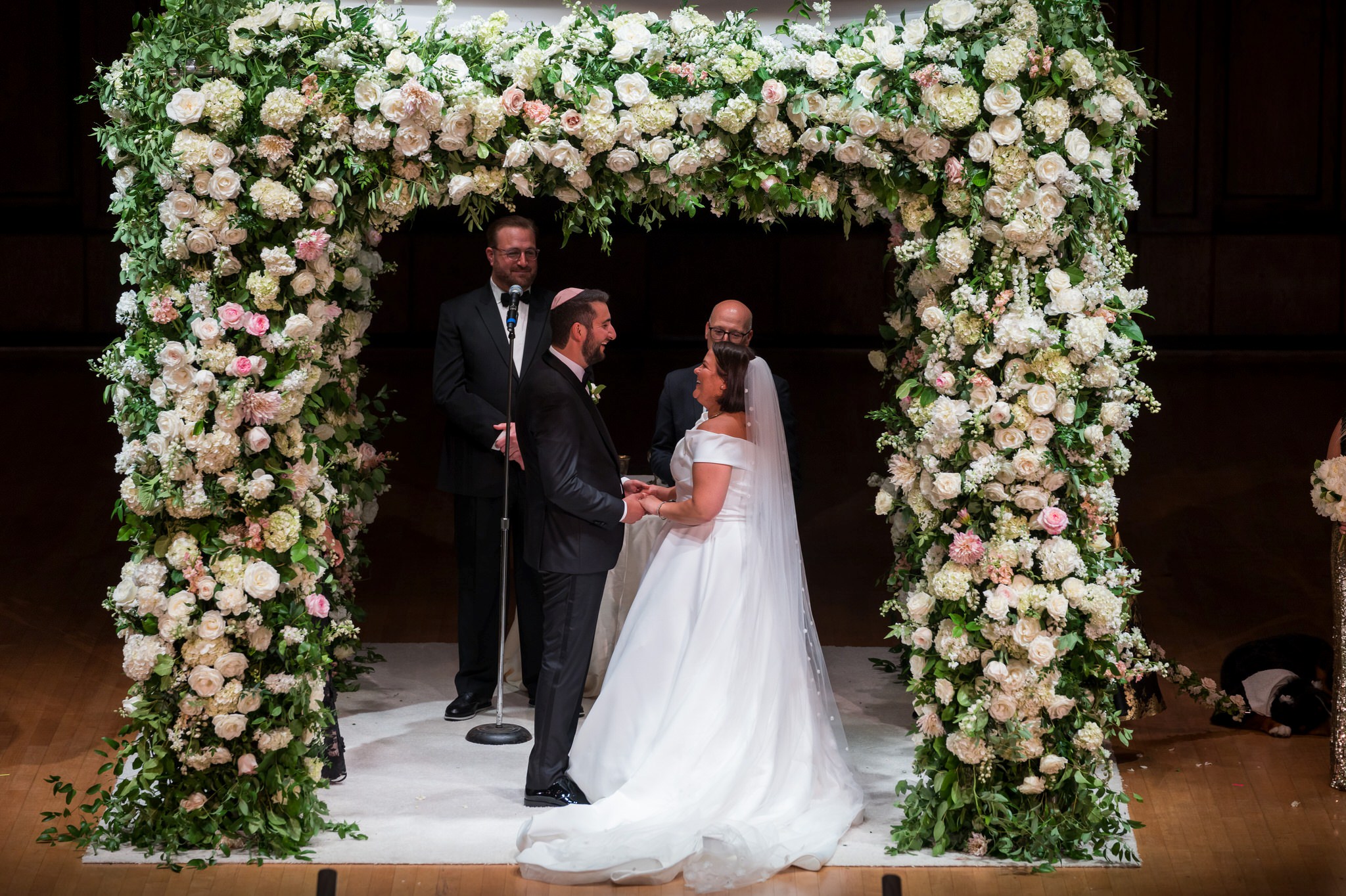 A jewish bride and groom smile, holding hands, and look at each other under a white chuppah at the Detroit Orchestra Hall wedding.  