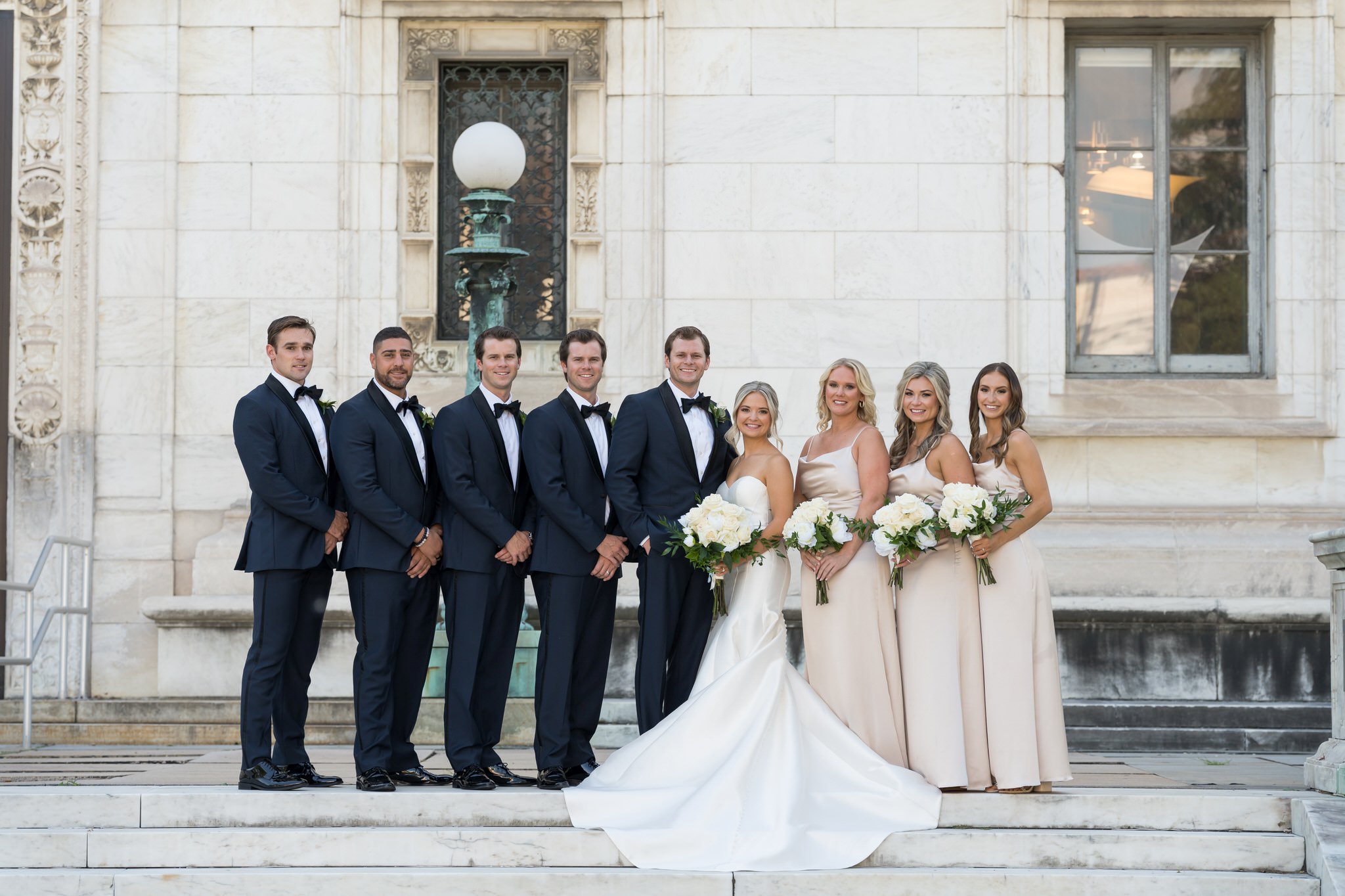 A bridal party wearing blue and black tuxedos and champagne dresses outside of the Detroit Public Library.  