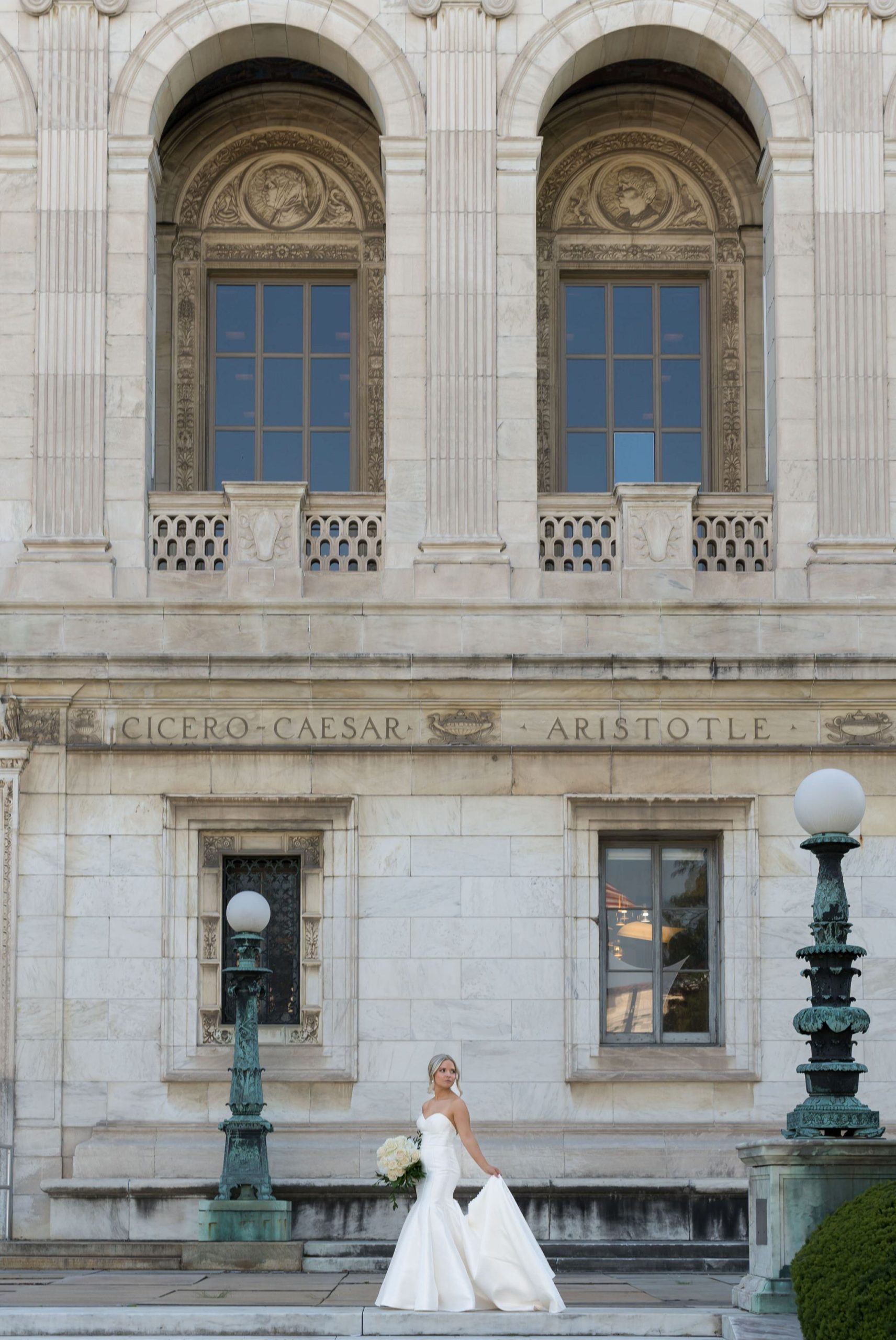 A bride holds the train of her dress and poses on the steps of the Detroit Public Library.  