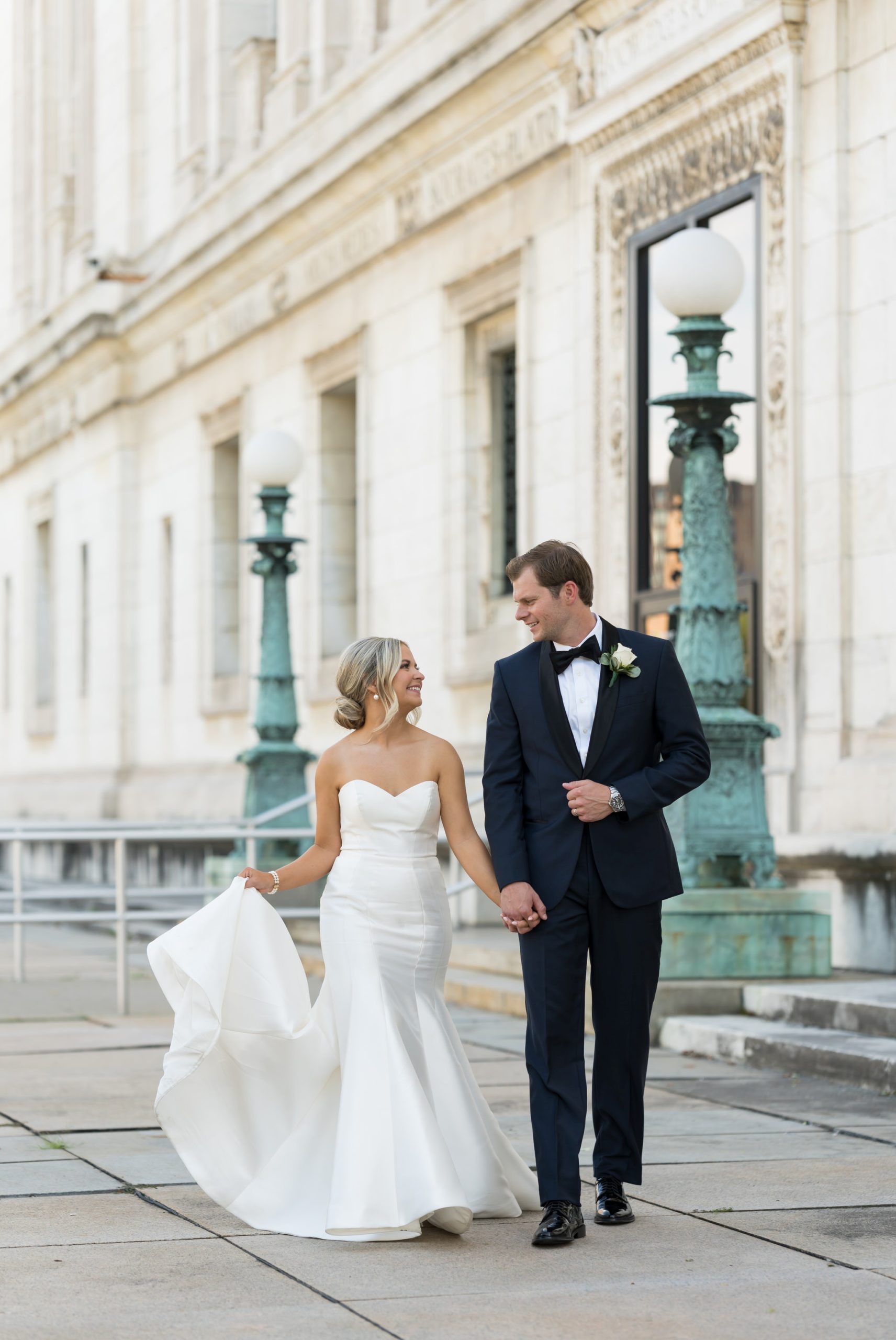 A bride, holding her dress, walks with her husband outside of the Detroit Public Library on their wedding day by Brian Weitzel Photography.