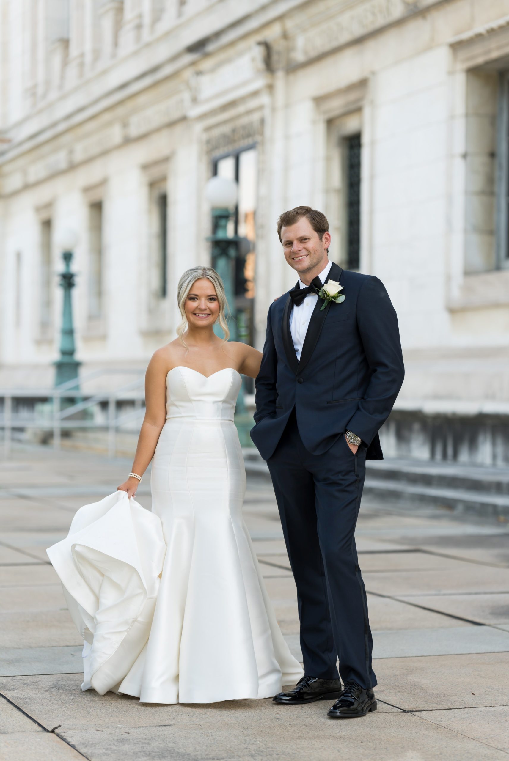 A bride, holding her dress, poses by her husband outside of the Detroit Public Library on their wedding day.  
