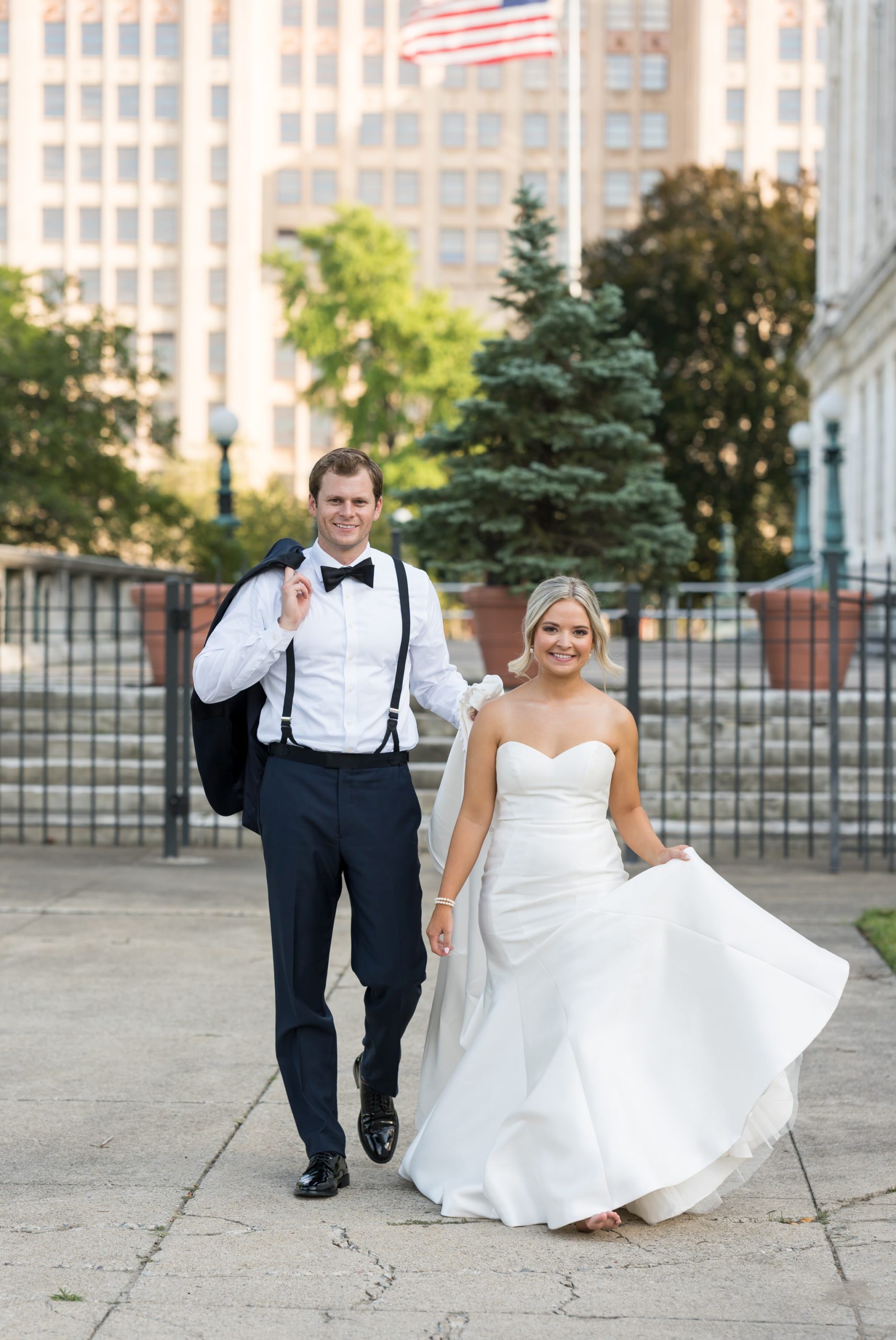 A groom, holding his tuxedo jacket over his shoulder, walks with his bride at the Detroit Public Library.  
