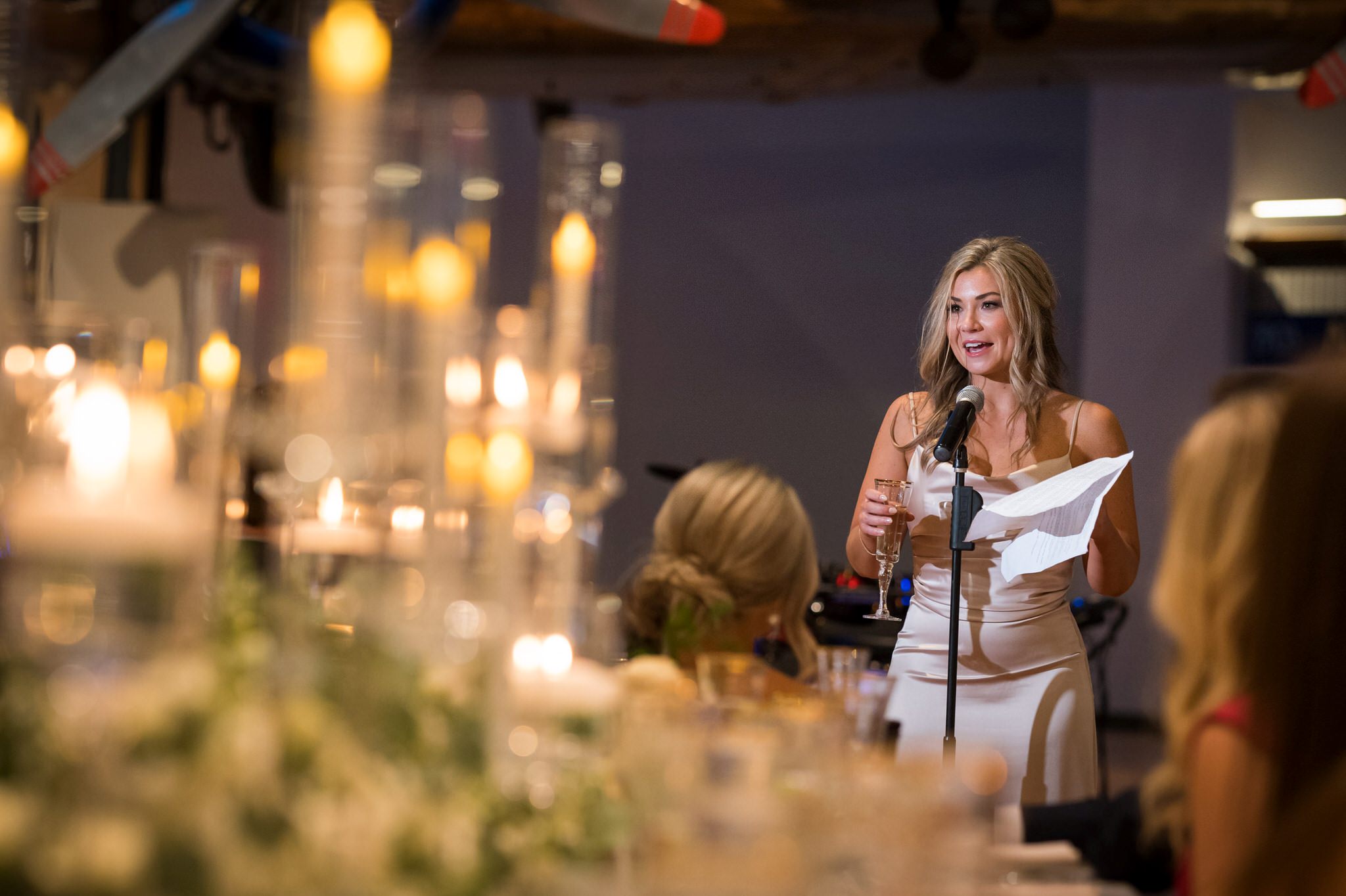 A bridesmaids, holding a champagne glass, gives a toast with blurry candles in the foreground at a Henry Ford Museum wedding. 