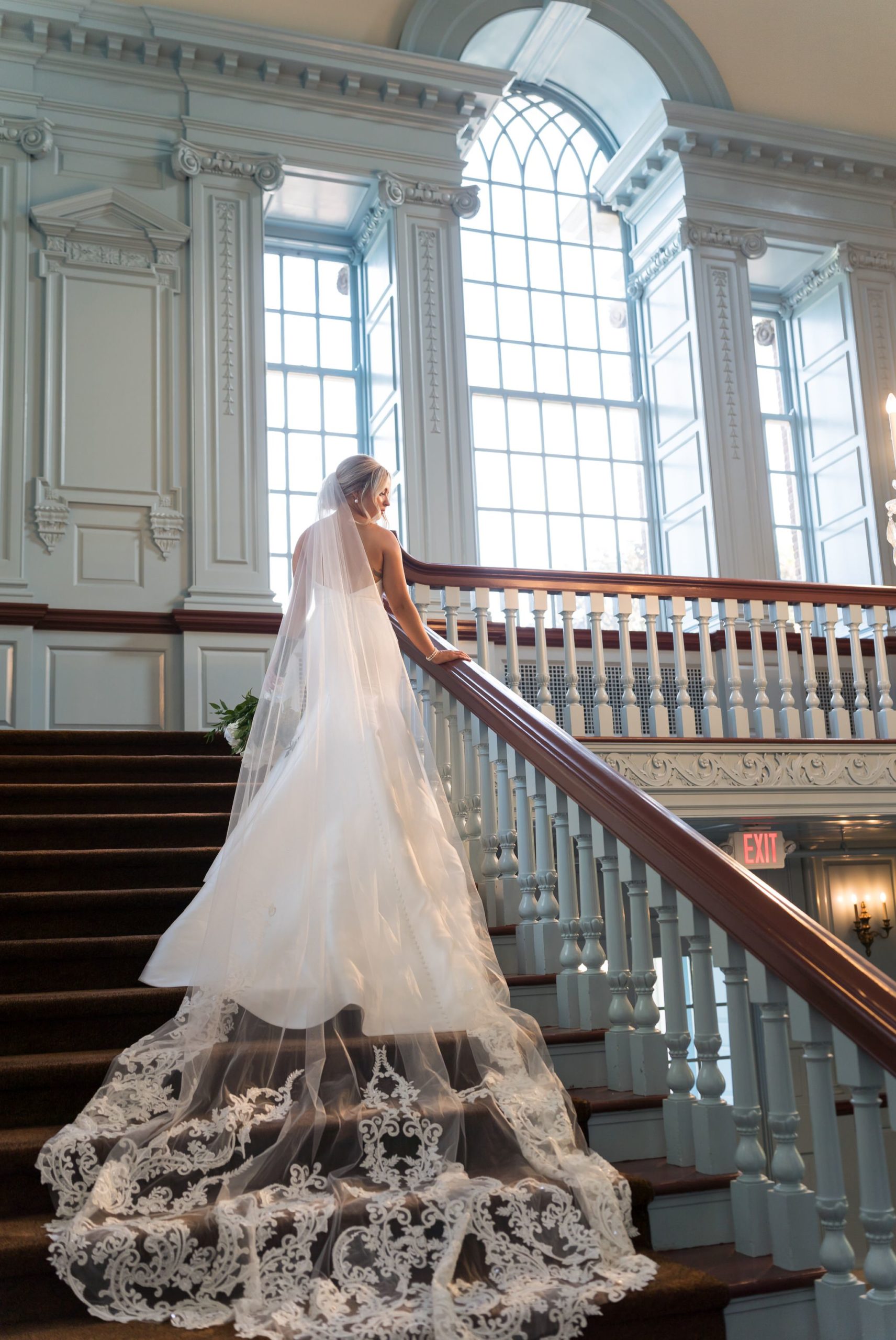 A bride holds the railing of a staircase  while her veil cascades behind her at her Henry Ford Museum wedding. 