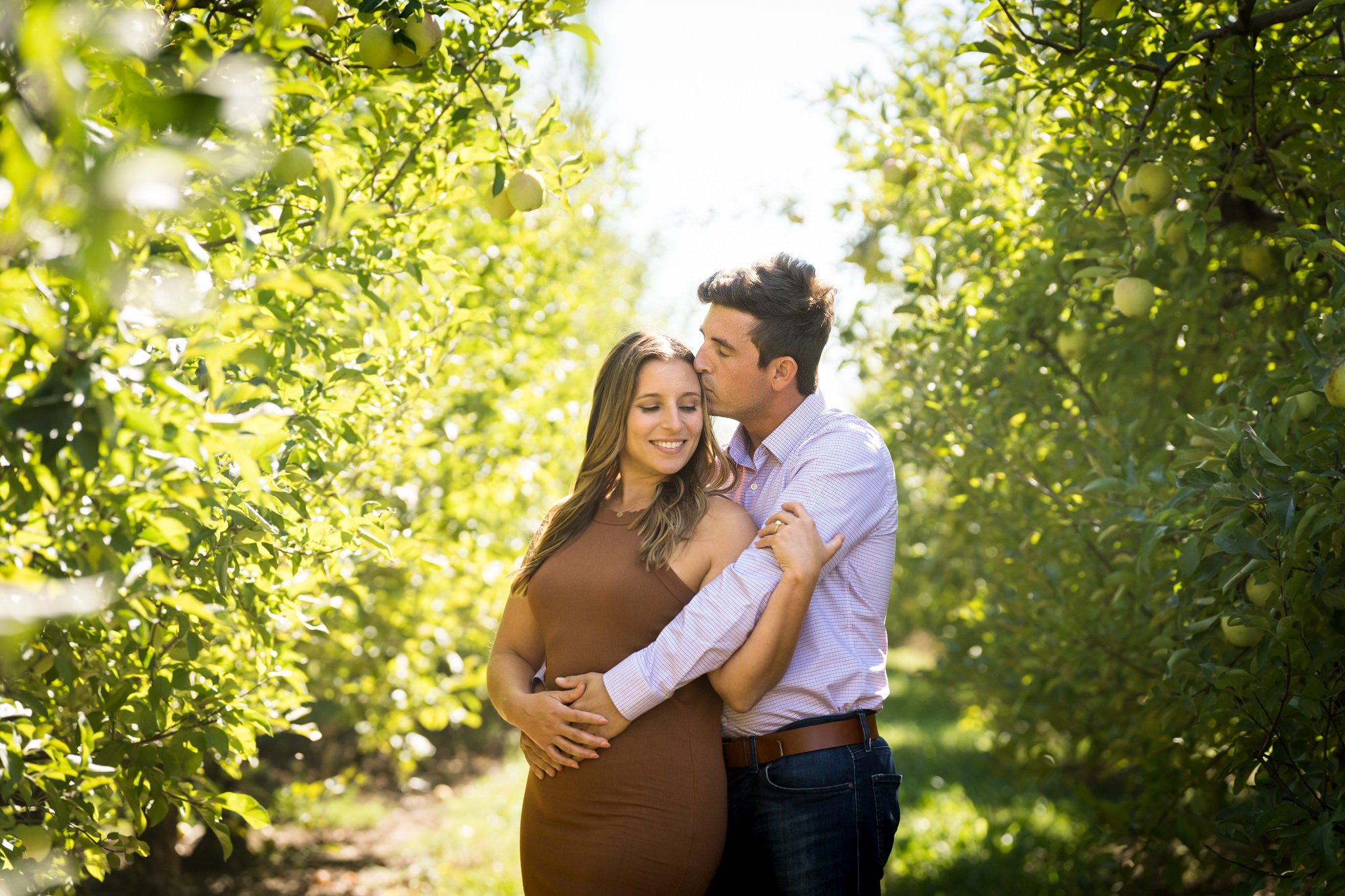 A man kissed the side of his fiance's head while hugging her, surrounded by apple trees at their Blake's Apple Orchard engagement session. 